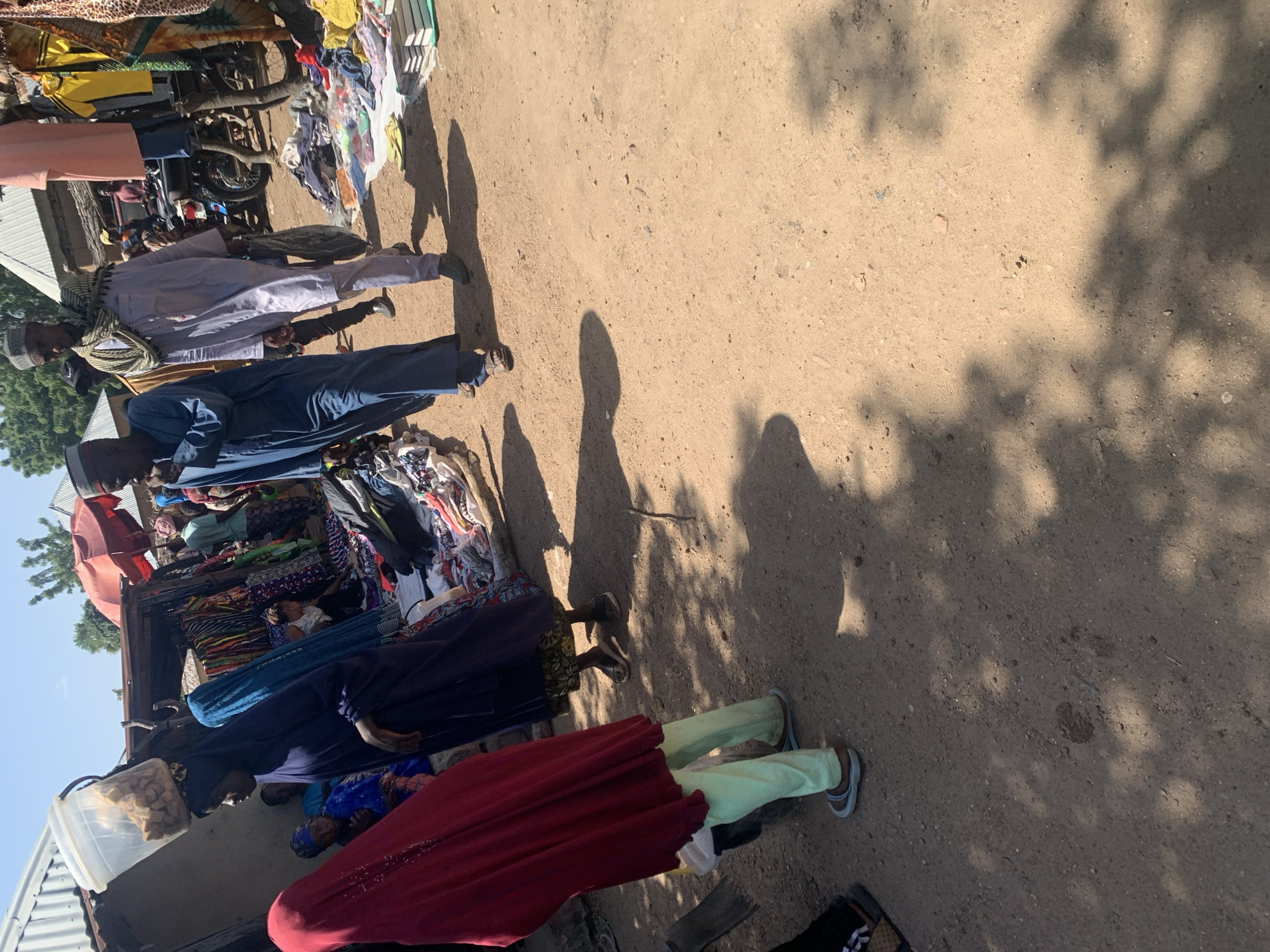 This is a medium shot, taken from eye-level, of a busy outdoor market scene in Boi A, Nigeria. The ground is a dusty, sandy dirt path, and the scene is dappled with shadows from overhead trees or structures.
In the foreground left, a person wearing a bright red hijab and light green pants walks away from the camera. To their right, a woman in a dark blue hijab and dress also walks away, carrying a bucket on her head filled with food items. Behind her, another woman in a blue headscarf is visible, looking towards the market.
The middle ground is dominated by market stalls. One stall has a variety of colorful textiles and clothing for sale, with mannequins or displays of garments. Another stall appears to be selling food or goods in containers. Several people are browsing or working at the stalls. A man in a light blue robe and cap walks towards the right of the frame, and another man in a similar light blue robe and a patterned scarf is walking next to him. A motorbike is parked near one of the stalls.
In the background, there are simple buildings with corrugated metal roofs, typical of the area. The sky is bright and clear, indicating a sunny day. The overall impression is one of vibrant activity and commerce in a rural Nigerian market.