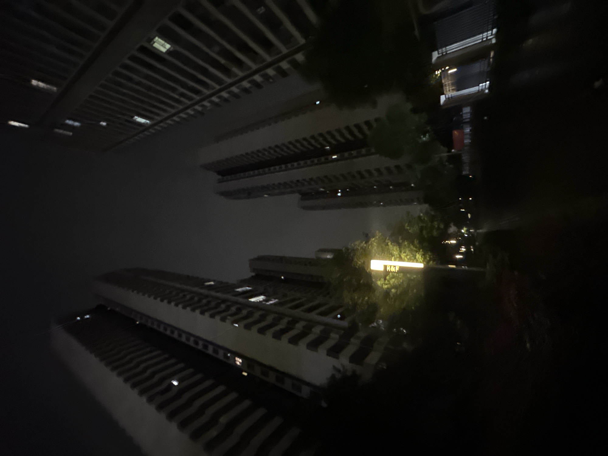 The image depicts a nighttime scene in the Chongqing High-tech Zone, China, characterized by an urban residential environment. Tall, dark high-rise buildings dominate the frame, angled upwards from a low perspective. On the left, two prominent residential structures exhibit numerous horizontal balconies or architectural features, with several windows emitting warm white or yellow light. Similar structures are visible on the right, also displaying illuminated windows. The sky is dark and overcast, lacking celestial features. In the mid-ground, a tall, vertical street lamp on the left emits bright yellow light, illuminating nearby green foliage. A dark pathway or road stretches into the distance, appearing slightly reflective. On the far right, a dark fence or gate structure with vertical elements is visible, and a faint red banner or flag-like object can be discerned in the distant mid-ground. The overall scene is devoid of human presence or overt actions, presenting a static nighttime cityscape under artificial illumination.
