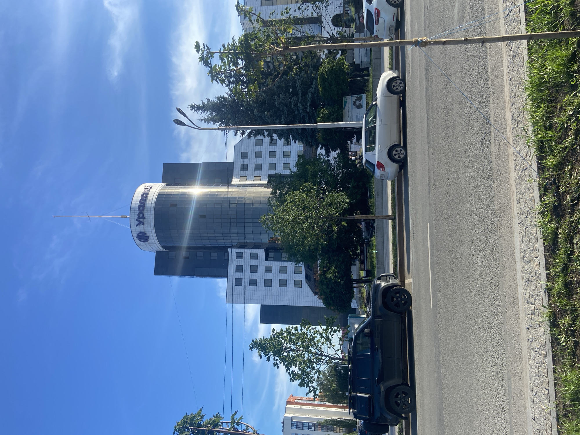 The image captures a view of a modern office building in Ufa, Russia, on a clear, sunny day. The building features a prominent cylindrical section topped with the logo "Уралсиб," which is a Russian financial corporation. The building is multifaceted, with a dark glass-enclosed top section and white paneling on its lower, angular sections. In front of the building, a street is visible with several cars parked and driving. A black SUV, possibly a Land Rover Defender, is parked on the left side of the road, and a white hatchback is seen further down the street. A tall street lamp with two lights stands near the building, alongside several mature evergreen trees and deciduous trees with green foliage. The sky is a vibrant blue with wispy white clouds. The foreground shows a patch of green grass bordering a paved sidewalk and the street. A thin wooden pole with blue string is positioned in the lower right corner of the frame.