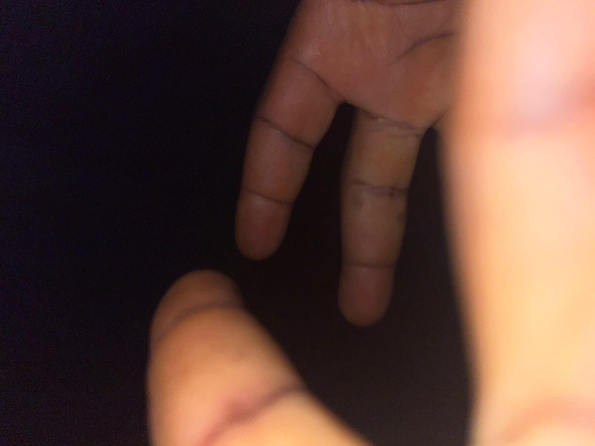 The image shows a close-up of two hands, with fingers in focus against a dark background. The skin tone is light brown, and there appear to be faint lines or marks on the fingers, possibly indicating scars or creases. The lighting is dim, with some areas of the hands being more illuminated than others, creating a soft glow. The context of Malejor, Ghana, is not visually represented in this image, as it solely focuses on the hands. There are no identifiable environmental features, buildings, or cityscapes visible.