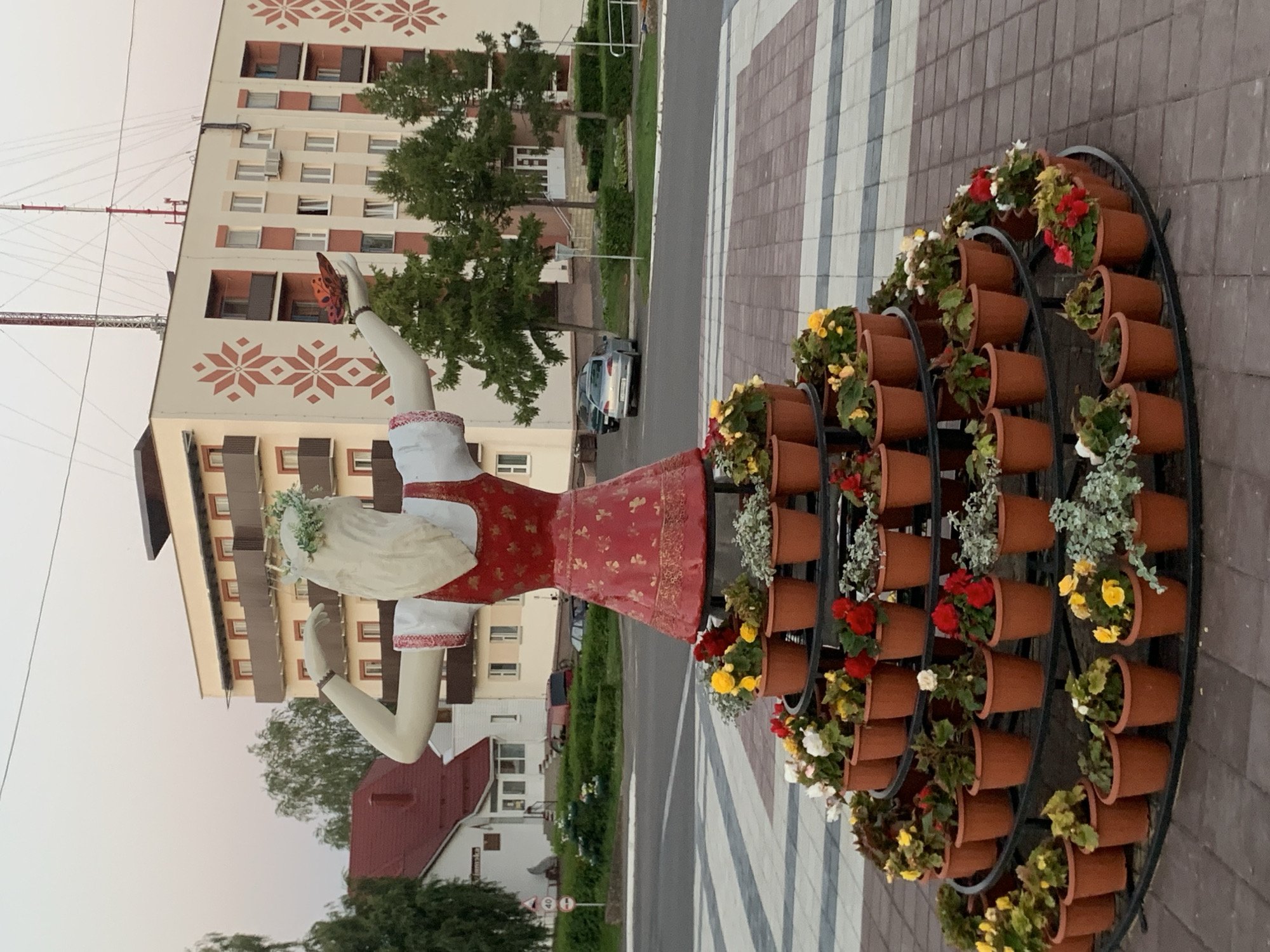 This image depicts a public display of flowers in front of a building in Kapyĺ, Belarus. The primary subject is a large, white statue of a woman wearing a red and white dress and a floral wreath. She is posed with one arm raised, holding an orange butterfly. The statue is adorned with numerous potted flowers arranged on tiered shelves around its base, featuring varieties of red, yellow, and white blossoms, as well as greenery. In the background, a multi-story building with a light-colored facade and dark balconies is visible, decorated with a red geometric pattern. Several trees, a street, and parked cars are also present, suggesting an urban environment. The sky is a soft, pale hue, indicating it might be early morning or late evening.