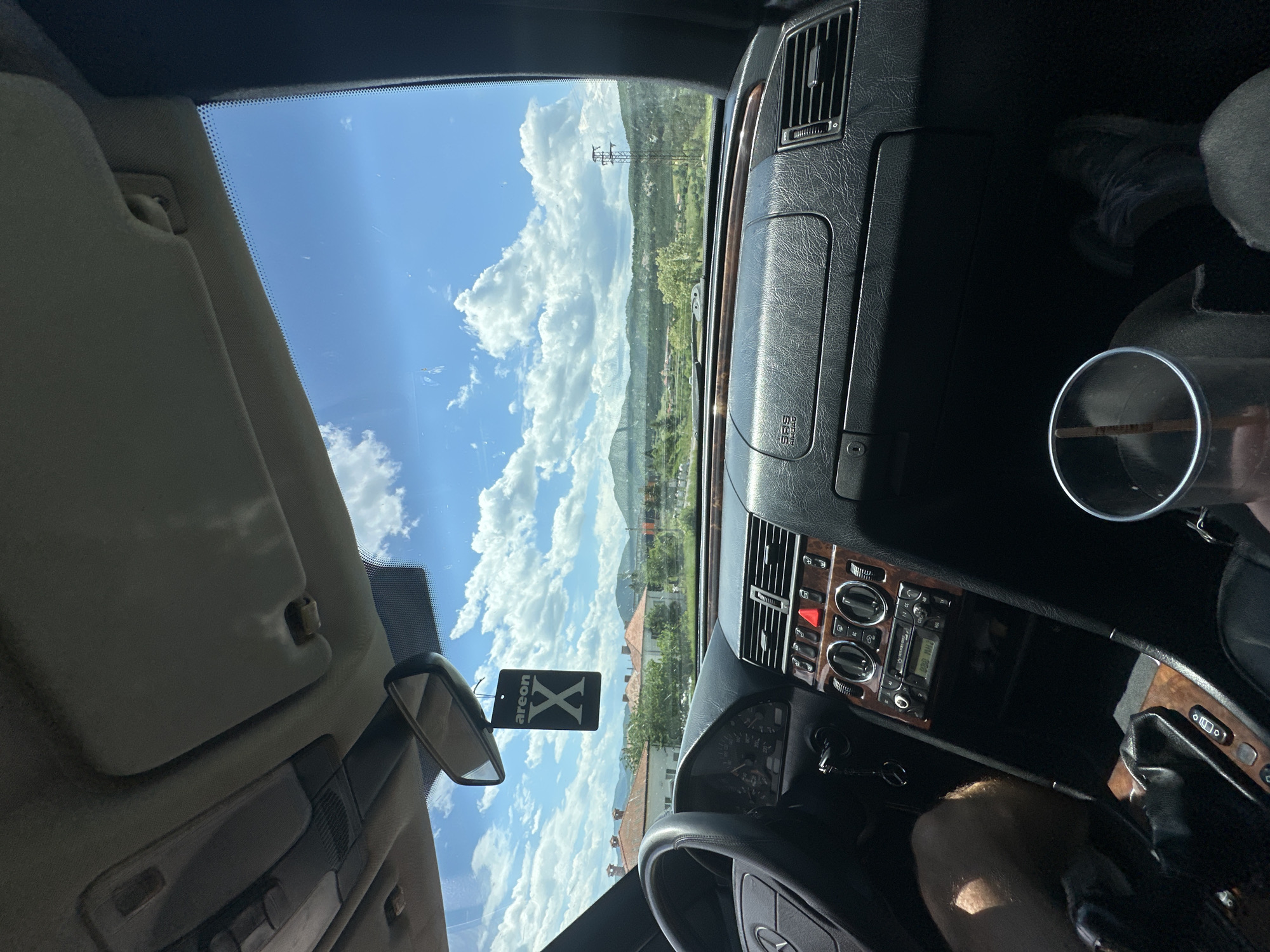The image shows the interior of a car, likely a Mercedes-Benz, looking out the windshield. The view from the windshield reveals a landscape with hills, a town, and a clear blue sky with scattered clouds. The presence of power lines suggests a rural or suburban environment. Inside the car, the dashboard, steering wheel, and part of the driver are visible. An "areon X" air freshener hangs from the rearview mirror. Based on the presence of Bulgarian writing on the radio, the scene is in Bulgaria, specifically near the town of Komuniga as claimed.