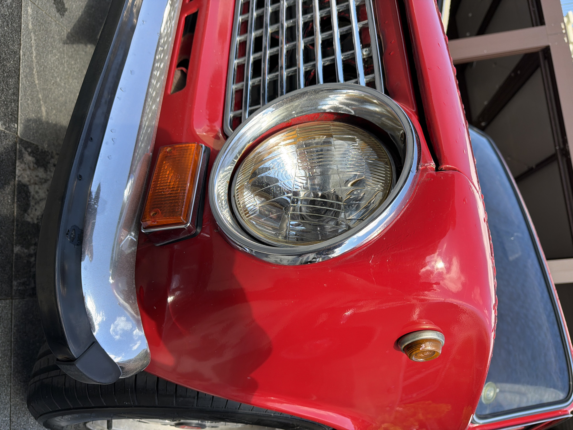 The image shows the front end of a red car. It features a chrome bumper with a black rubber trim, a chrome grille, a round headlight with a chrome bezel, and an amber turn signal indicator. The car appears to be an older model. The background suggests an outdoor urban setting. There is no clear indication in the image to confirm that the location is Moscow, Russia.