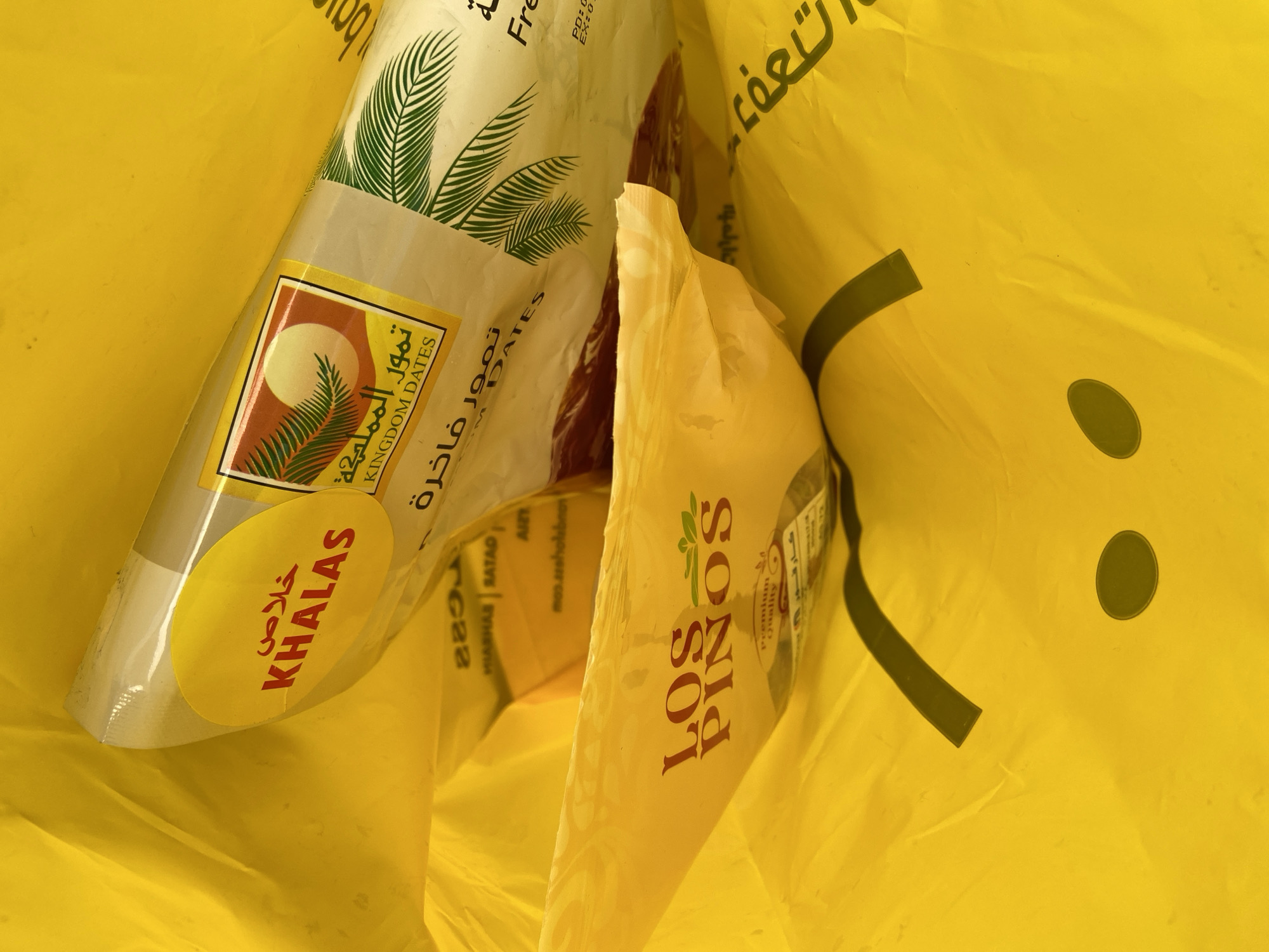 The image provides an overhead view looking directly into a bright yellow plastic shopping bag, which is crumpled and takes up most of the frame. Inside the bag are two distinct product packages.
One package is a clear plastic container of dates, prominently featuring a label with a desert scene of a sun and a palm tree. The label reads "تمور المملكة" (Kingdom Dates) in Arabic, with "KINGDOM DATES" in English below. A yellow oval sticker on this package clearly states "خلاص" (KHALAS), identifying the variety of dates.
The second package, partially visible and somewhat obscured by the folds of the yellow bag, shows a label with the brand name "LOS PIÑOS" and "Premium Quality" written beneath it.
Various fragments of text are visible on the plastic bag and packages. On the yellow bag, partially visible Arabic characters include "تعة" (possibly part of "enjoyment"). English fragments like "Fre" (likely "Fresh"), "PD:" (Production Date), and "EX:" (Expiry Date) can also be seen. Other obscured text includes "MIARHAS RATAD", "AIZY", "mos.azaholebnos", "كارفي", and "والمياه".
The overall scene depicts purchased items, presumably groceries or snacks, contained within a plastic carrier bag. The lighting is soft and diffused, without strong shadows, suggesting an indoor setting or indirect light. While nothing in the image explicitly indicates the location, the items are situated in Budva, Montenegro, based on the provided context. No specific activity is taking place, rather it's a still life composition of the bag's contents.
