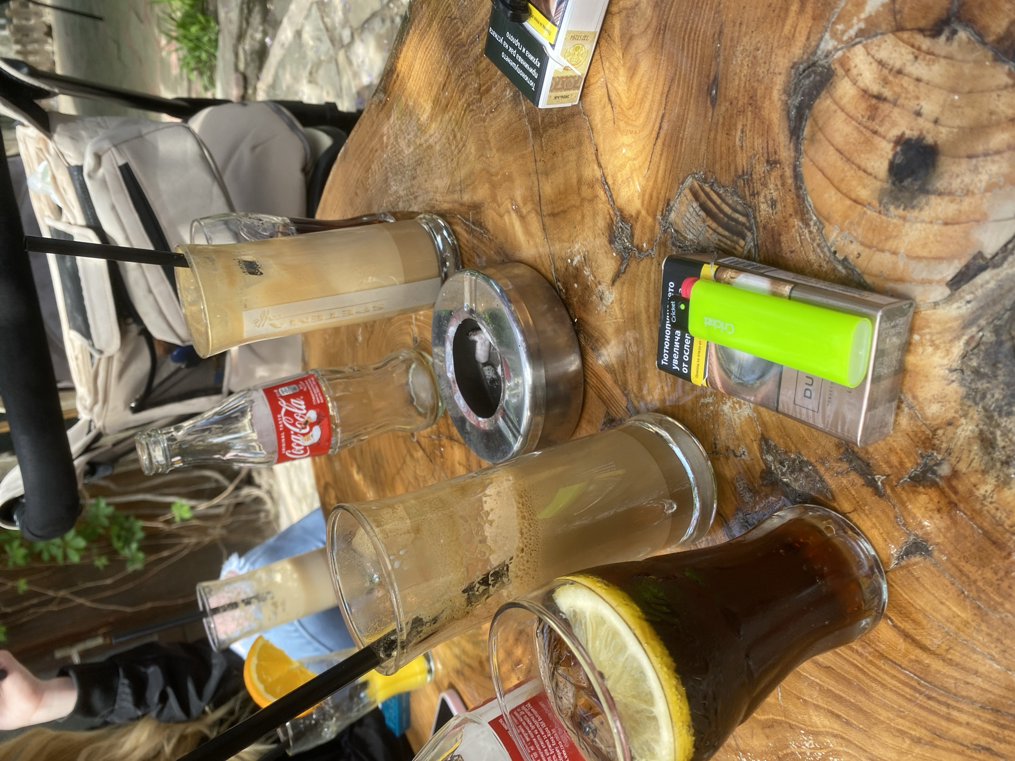 The image captures a scene at a cafe, likely outdoors given the wooden table and partial view of greenery. Several drinks are present, including two iced coffee drinks with straws, a Coca-Cola bottle, and another drink, possibly iced tea, with a lemon slice. The table also contains an ashtray, a pack of cigarettes, and a lighter. The presence of cigarette paraphernalia suggests that smoking is permitted in the cafe. The visible text on the cigarette pack is in Bulgarian. The text on the Coca-Cola bottle confirms it is from Coca-Cola HBC Bulgaria. The word 'Caffe' can be seen on one of the glasses. Without further information, determining if the location is specifically in Varna, Bulgaria, is not possible.