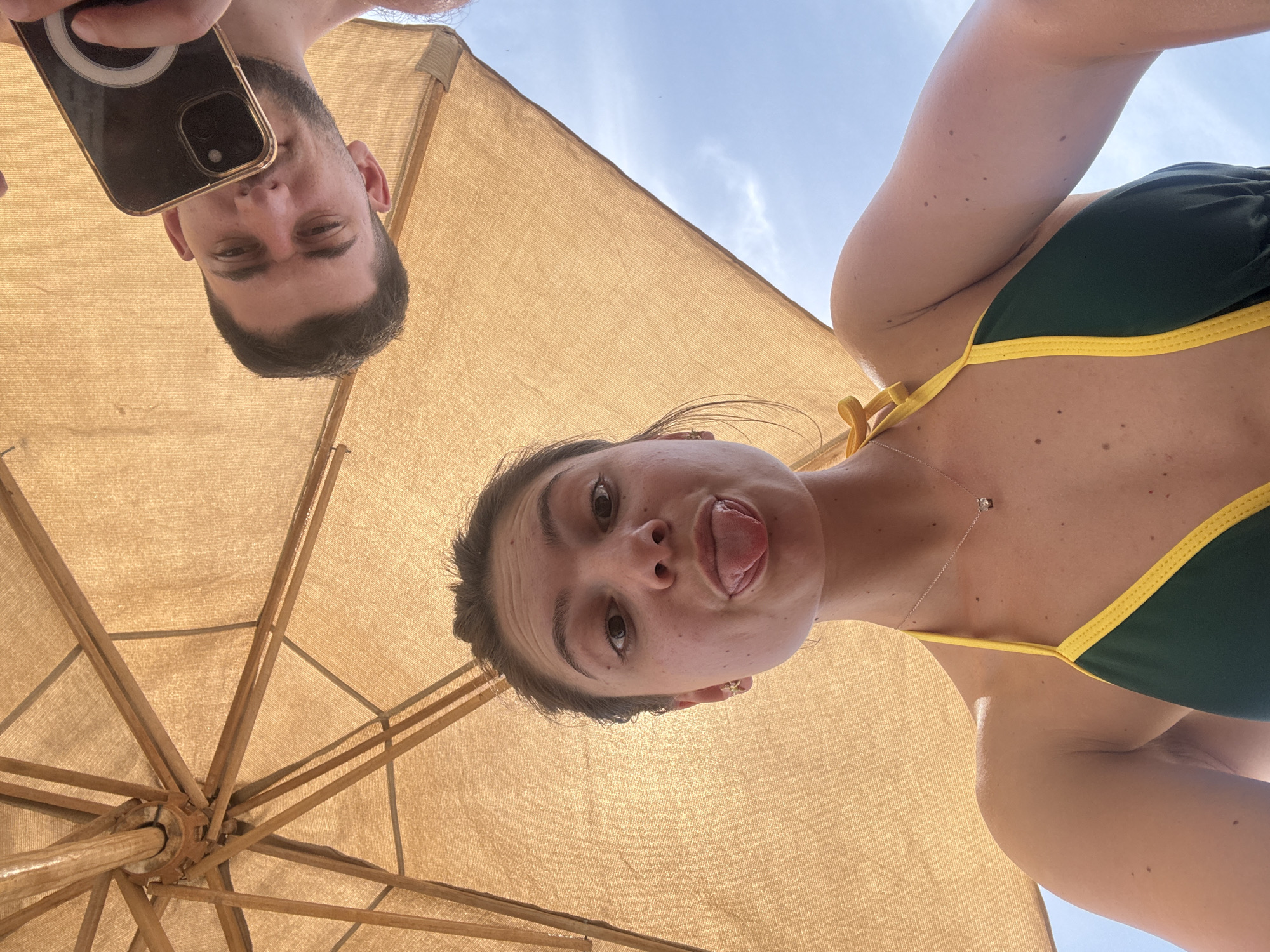 Two individuals are positioned under a tan-colored umbrella. A male is holding a smartphone, his face visible above the umbrella's edge. A female is beside him, wearing a green and yellow bikini, and she is sticking her tongue out towards the camera. The umbrella's wooden structure and fabric are visible, along with a patch of blue sky. The location is Unknown, Unknown.