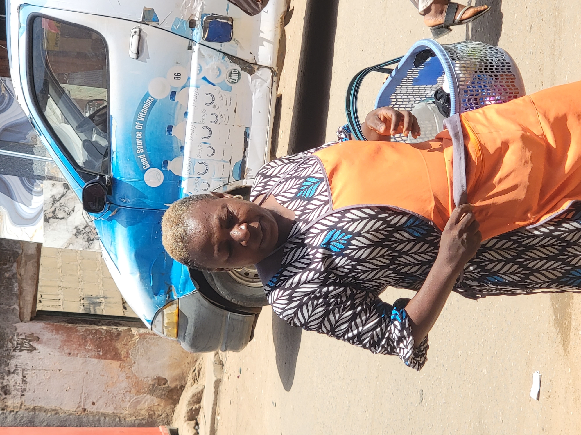 The image captures a person standing beside a three-wheeled vehicle on a sunny day in what appears to be a street or open area in Maiburiji, Nigeria.
The main subject is a person with dark skin and short, light-colored hair, looking directly at the camera with a calm, neutral expression. They are dressed in a patterned garment featuring black, white, and blue leaf motifs, covered by a plain orange apron. Their hands are holding the strap of a light blue and white mesh basket, which is positioned at their hip, likely containing goods.
Behind the person and to their left is a light blue and white three-wheeled vehicle, resembling an auto-rickshaw or Keke Napep, commonly used for transportation or deliveries. The vehicle is extensively branded with promotional text and graphics. Visible text includes "Good Source Of Vitamins", the number "86", "fresh%", and "Zinc". The graphics depict numerous small bottles, strongly suggesting the vehicle is associated with a beverage or dairy product, possibly milk.
The scene takes place outdoors on a light, sandy or dusty ground. The presence of strong sunlight and distinct shadows cast by the person and the vehicle indicates it is daytime, likely midday or early afternoon. In the background, there are hints of structures, including a rough-textured wall and some corrugated metal sheeting. A foot wearing a dark sandal is partially visible on the right side of the frame, suggesting another individual is nearby. The overall impression is a candid moment from daily life or commerce in a West African setting.