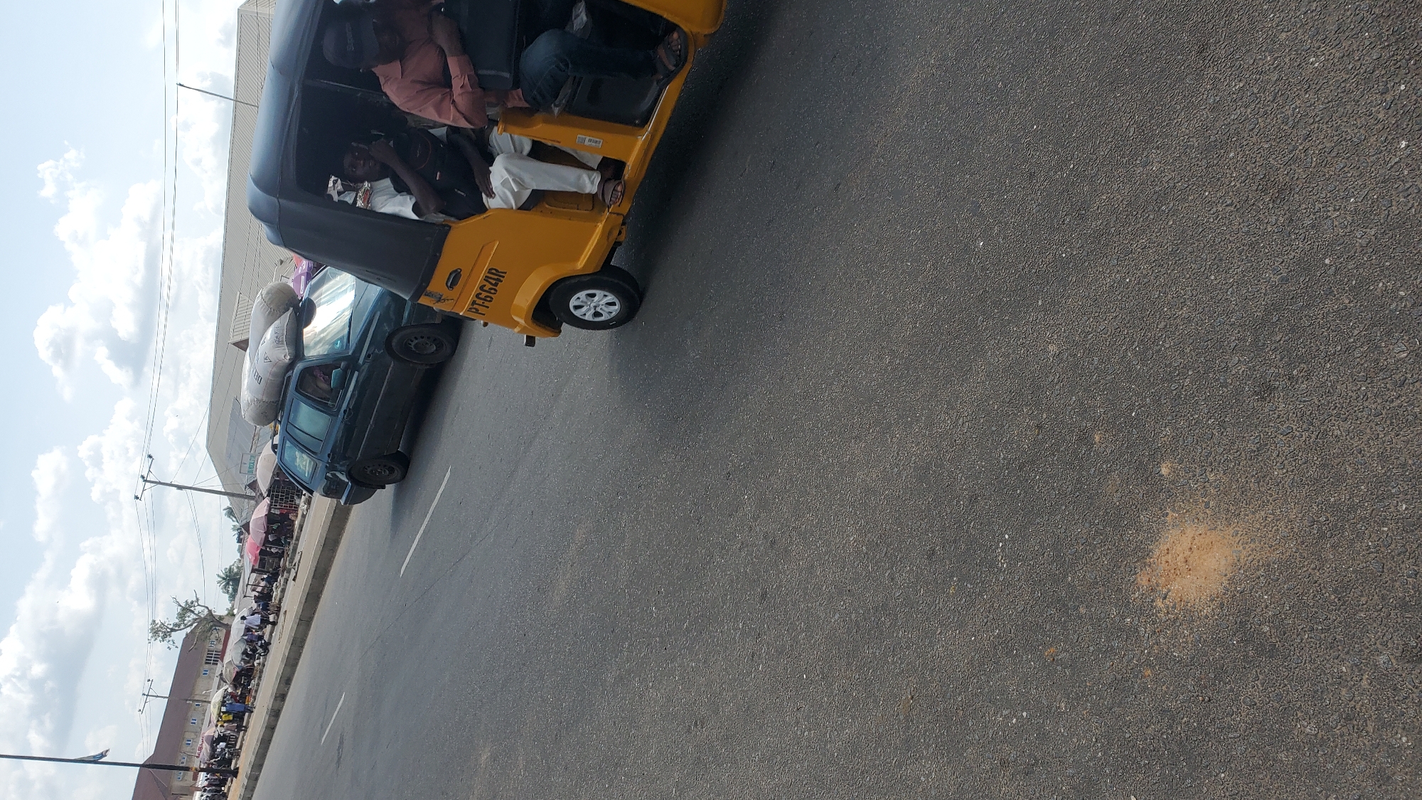 This image shows a street scene in Jalingo, Nigeria, with a yellow auto-rickshaw in the foreground and a dark green station wagon in the background. The auto-rickshaw is carrying three passengers, two in the back and one driver in the front. The station wagon has two large sacks of what appear to be grain or feed loaded on its roof rack, and one person is visible inside the vehicle. The street is paved with asphalt, and there are buildings and market stalls lining the side of the road. The sky is partly cloudy. The overall impression is of a busy, everyday scene in a Nigerian city.