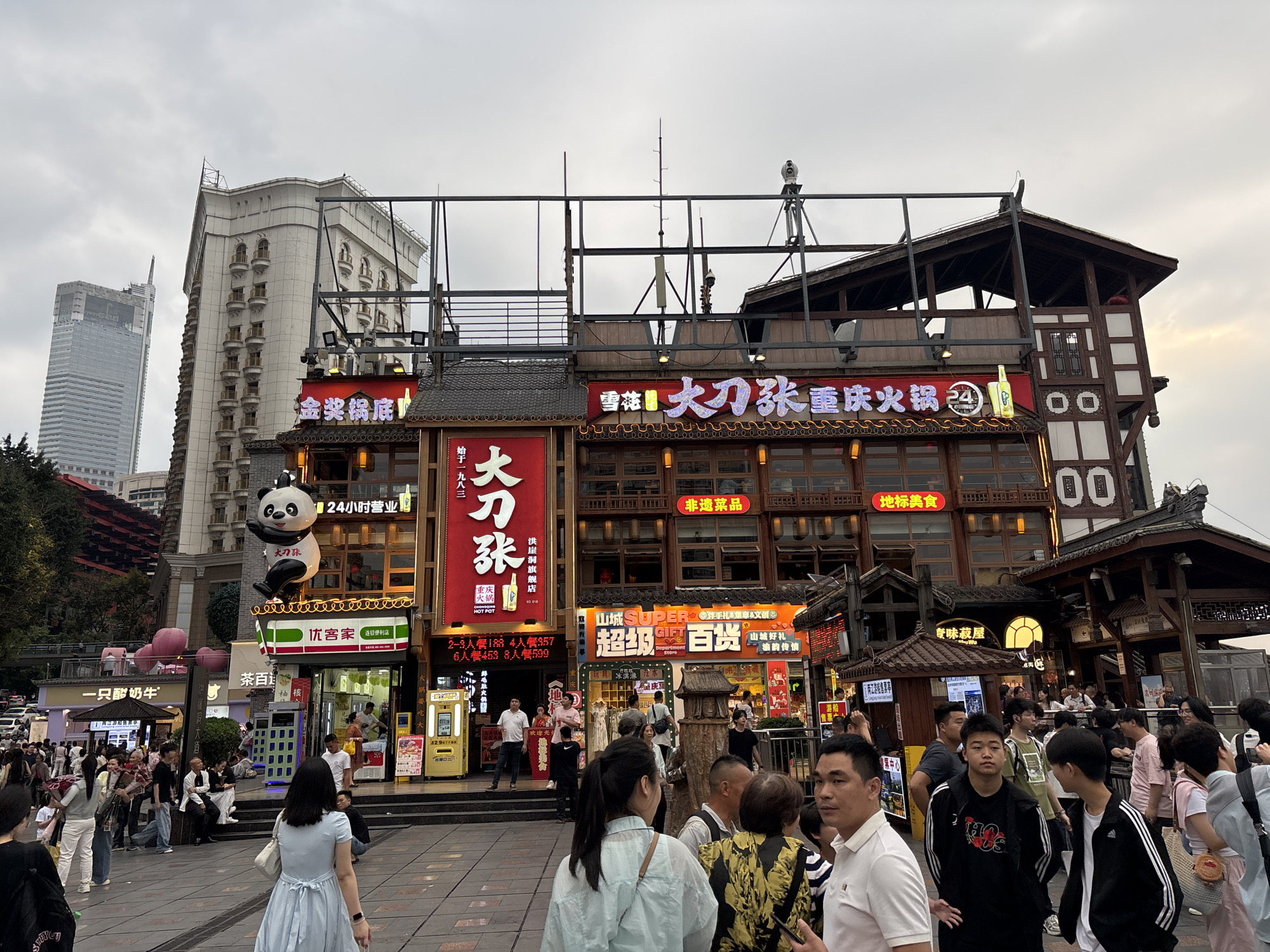 The photograph captures a bustling street scene in the Yuzhong District of Chongqing, China. The street is filled with pedestrians, many wearing casual attire. The backdrop features traditional Chinese architecture interspersed with modern buildings. A large panda statue adorns the storefront of a convenience store, "Youkejia," which is identified by its green and white signage. Several restaurants are present, displaying large Chinese characters and neon lighting. The building's rooftops are adorned with metal supports. The sky is overcast, casting soft light. Banners and signs advertise hot pot restaurants and department stores.