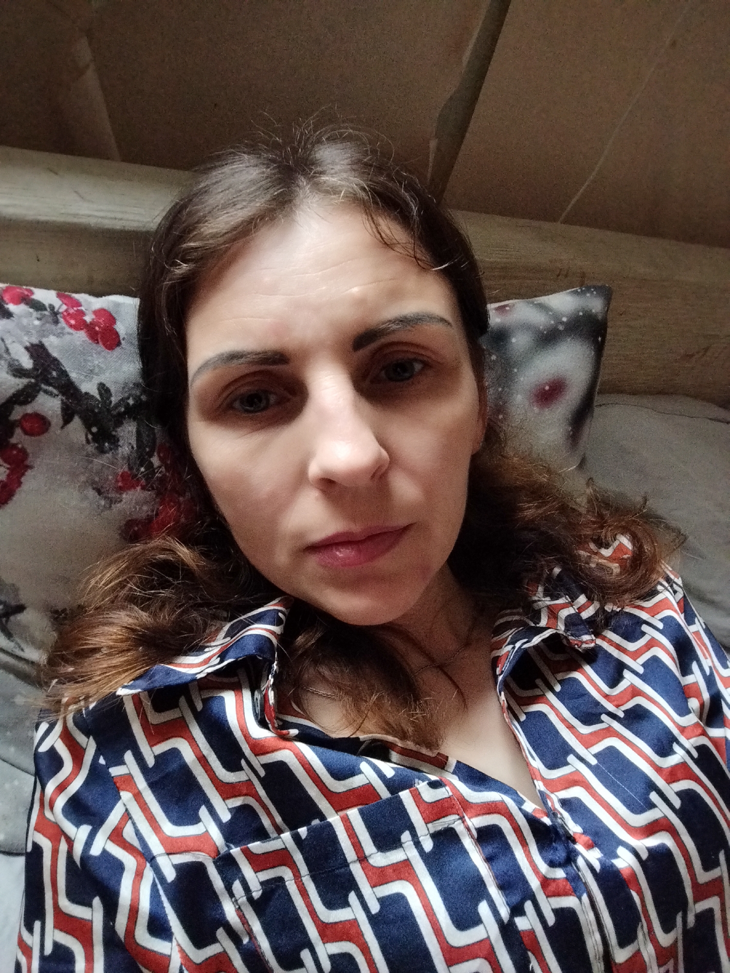 A woman with brown hair and blue eyes is lying on a bed and looking at the camera. She is wearing a shirt with a geometric pattern of blue, white, and red lines. The bed has pillows with a floral pattern on the left and a space-themed pattern on the right. The background appears to be a wooden surface. The image was taken in Vinnytsia, Ukraine.