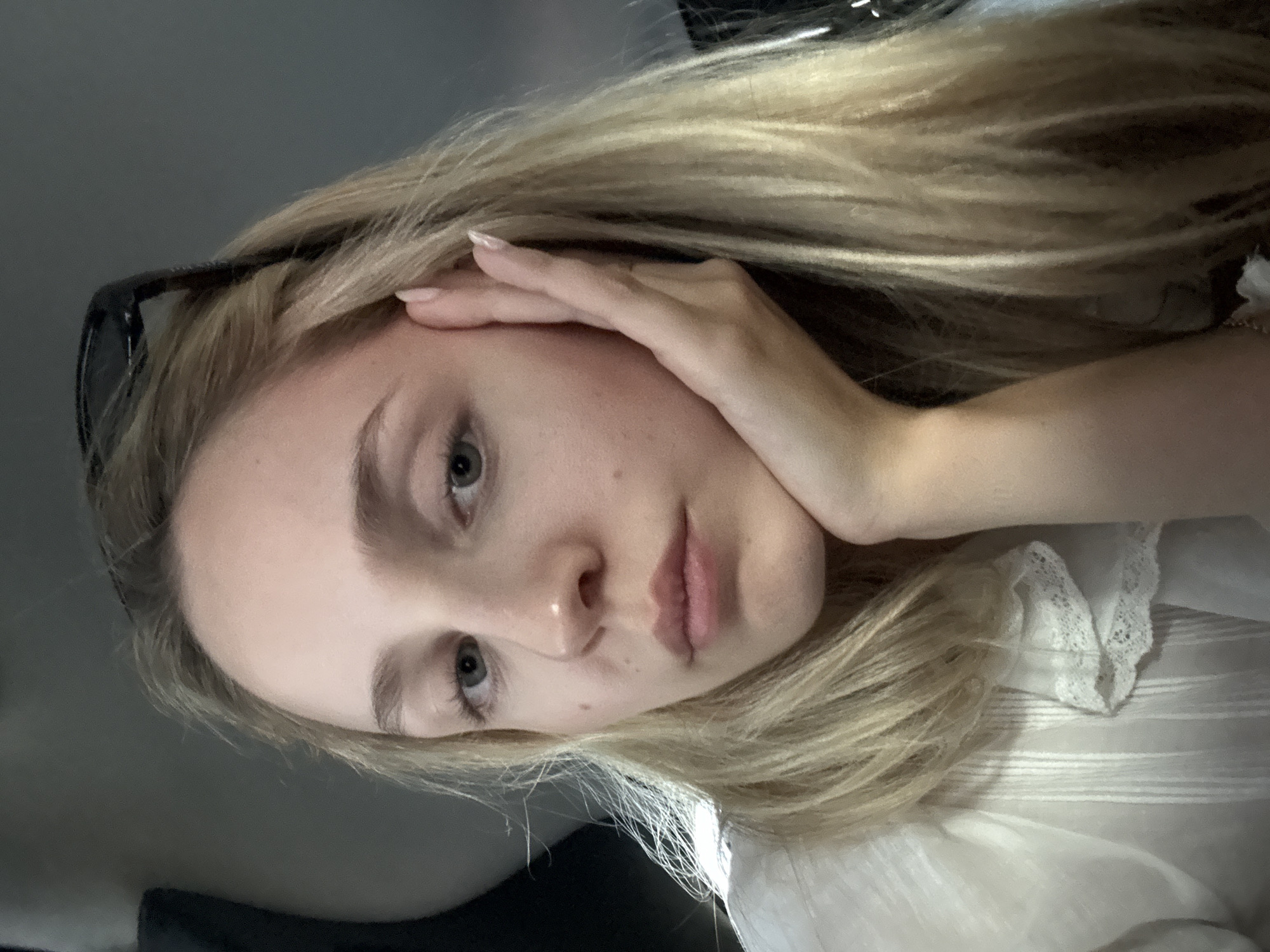 A young Caucasian woman with blonde hair is pictured indoors, resting her head on her hand and looking towards the camera. She is wearing a white shirt with lace trim and has sunglasses perched on top of her head. The background is dark and out of focus, suggesting an indoor setting with limited lighting. There are no visible landmarks or indicators to suggest a specific location within New York City, USA.
