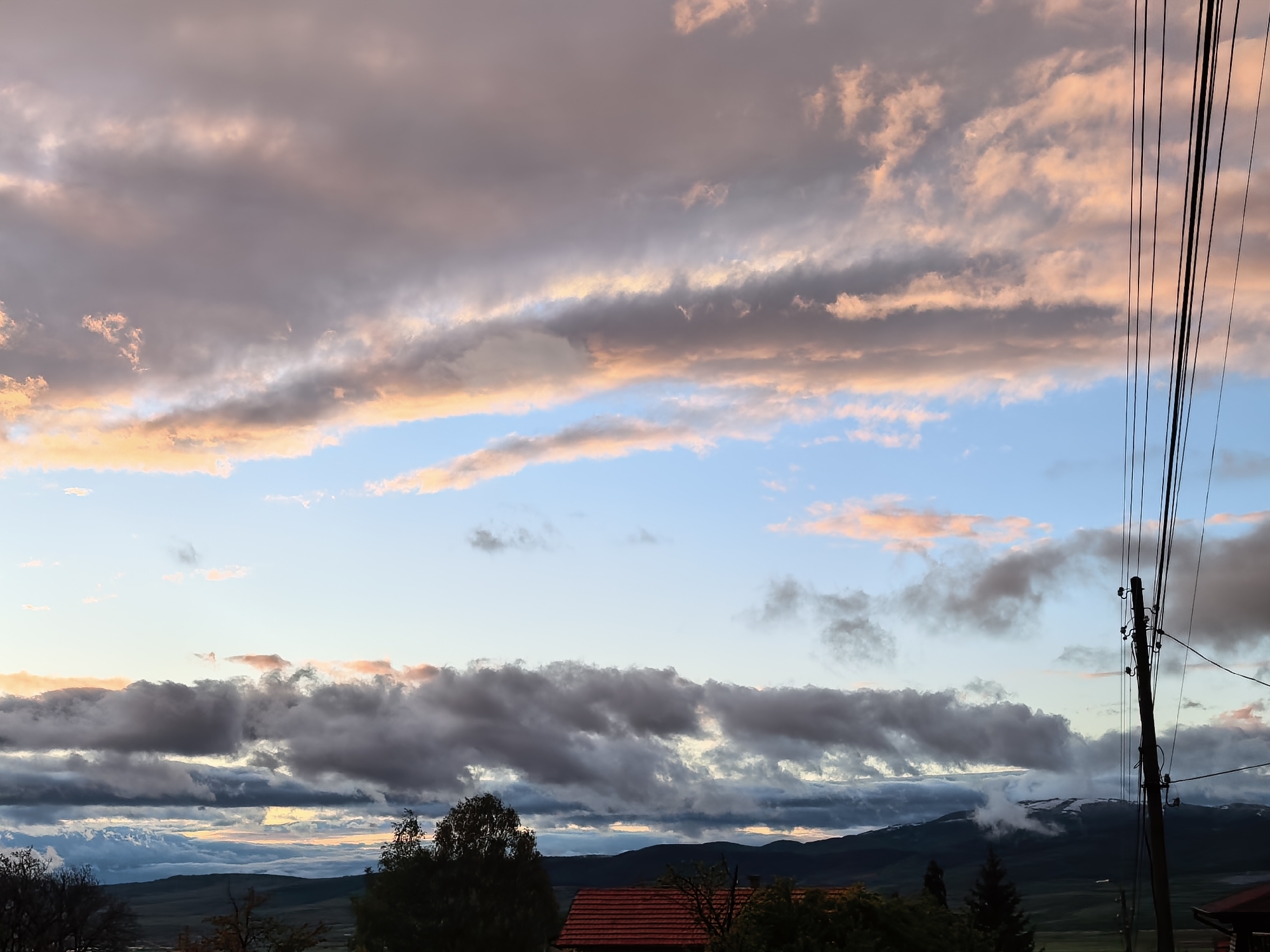 The image presents a scenic landscape dominated by a dramatic and colorful sky, likely captured during either sunrise or sunset over Sapareva Banya, Bulgaria. The sky is a prominent feature, filled with layers of clouds. The upper sections display expansive, diffuse grey clouds interspersed with clear blue sky, many of which are illuminated with soft, warm tones of orange, pink, and peach, suggesting the golden hour. Below these, a thick band of darker, denser grey clouds stretches across the middle of the frame, creating a striking contrast with the lighter hues above and below.
In the lower half of the image, distant mountains are visible, their dark silhouettes appearing rugged and undulating. Some of these peaks show lighter patches, possibly indicating snow or distinct rock formations. A valley or rolling hills can be discerned in the middle ground. The immediate foreground features dark silhouettes of trees and foliage. Towards the bottom right, the red-tiled roof of a building is partially visible amongst the dark trees.
A significant man-made element is a tall, dark utility pole positioned on the far right, extending vertically upwards. Numerous black power lines run diagonally and horizontally from this pole, cutting across the sky and partially obscuring the cloud formations. The scene evokes a sense of tranquil grandeur, characteristic of a mountainous region at dawn or dusk, with no visible text in the image.