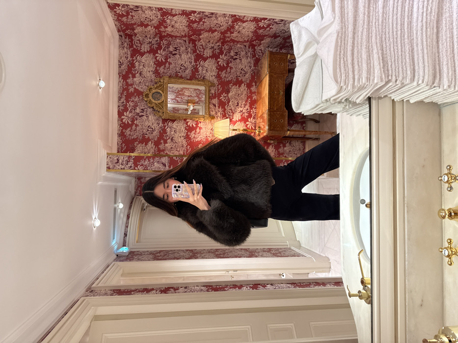 A woman in a dark furry jacket and black pants poses for a selfie in a luxurious bathroom. Red toile wallpaper adorns the walls, complemented by a framed mirror and a wooden vanity. A white marble bathtub with gold fixtures is visible on the right. A stack of white towels sits above the bathtub. The scene suggests an opulent hotel or residence.