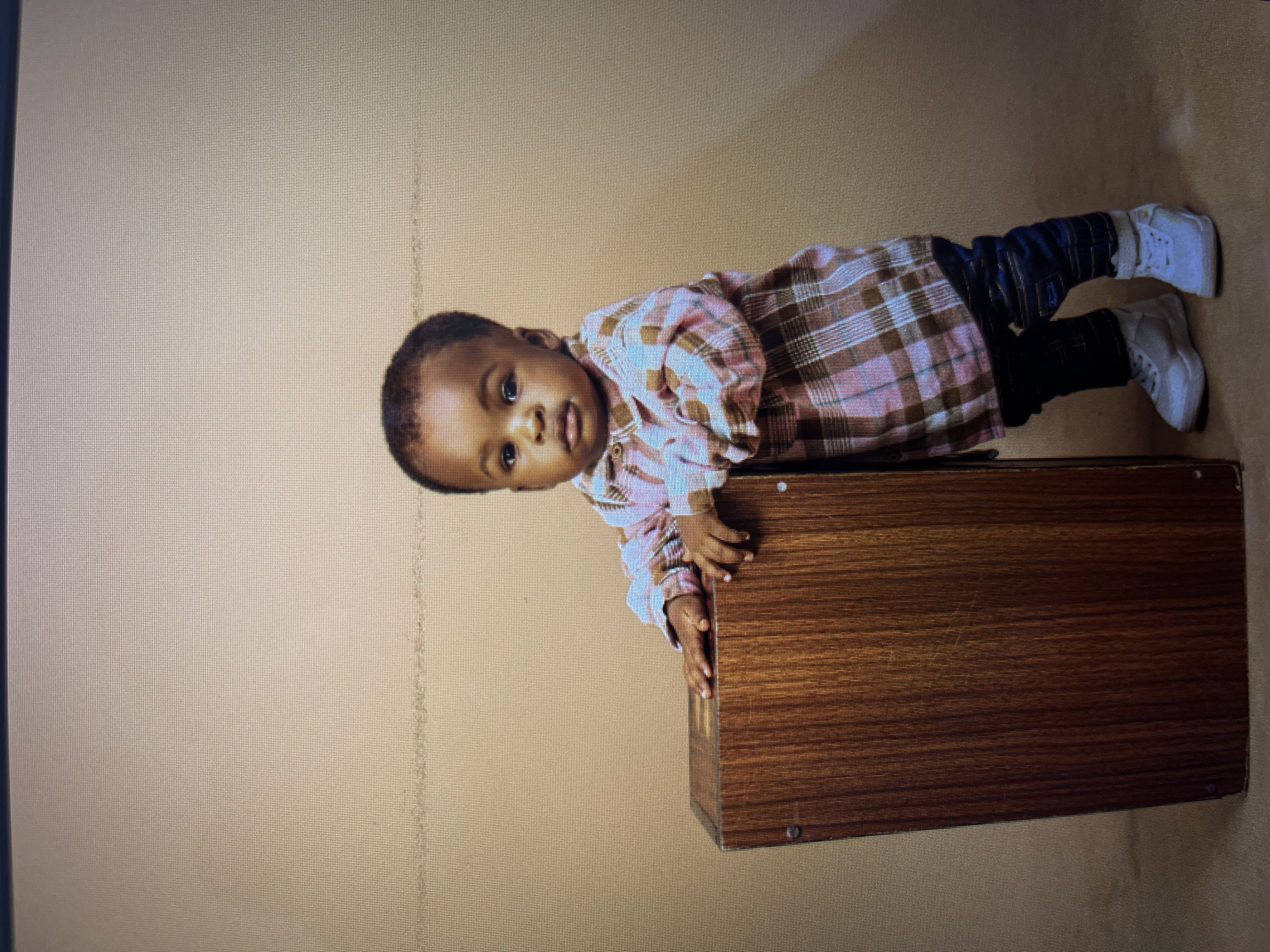 A young Black child, appearing to be between one and two years old, stands with their left side leaning against a dark wood, rectangular object, possibly a small table or a musical instrument like a cajon. The child is wearing a pink and brown plaid collared shirt, dark blue jeans, and white sneakers. Their hands rest on the top surface of the wooden object. The background is a plain, light brown studio backdrop. There is a faint horizontal line across the upper portion of the backdrop. The lighting is even, with no harsh shadows. The child's gaze is directed towards the camera. The scene appears to be a studio portrait setting, with no indications of a specific location in Gura Top, Nigeria.