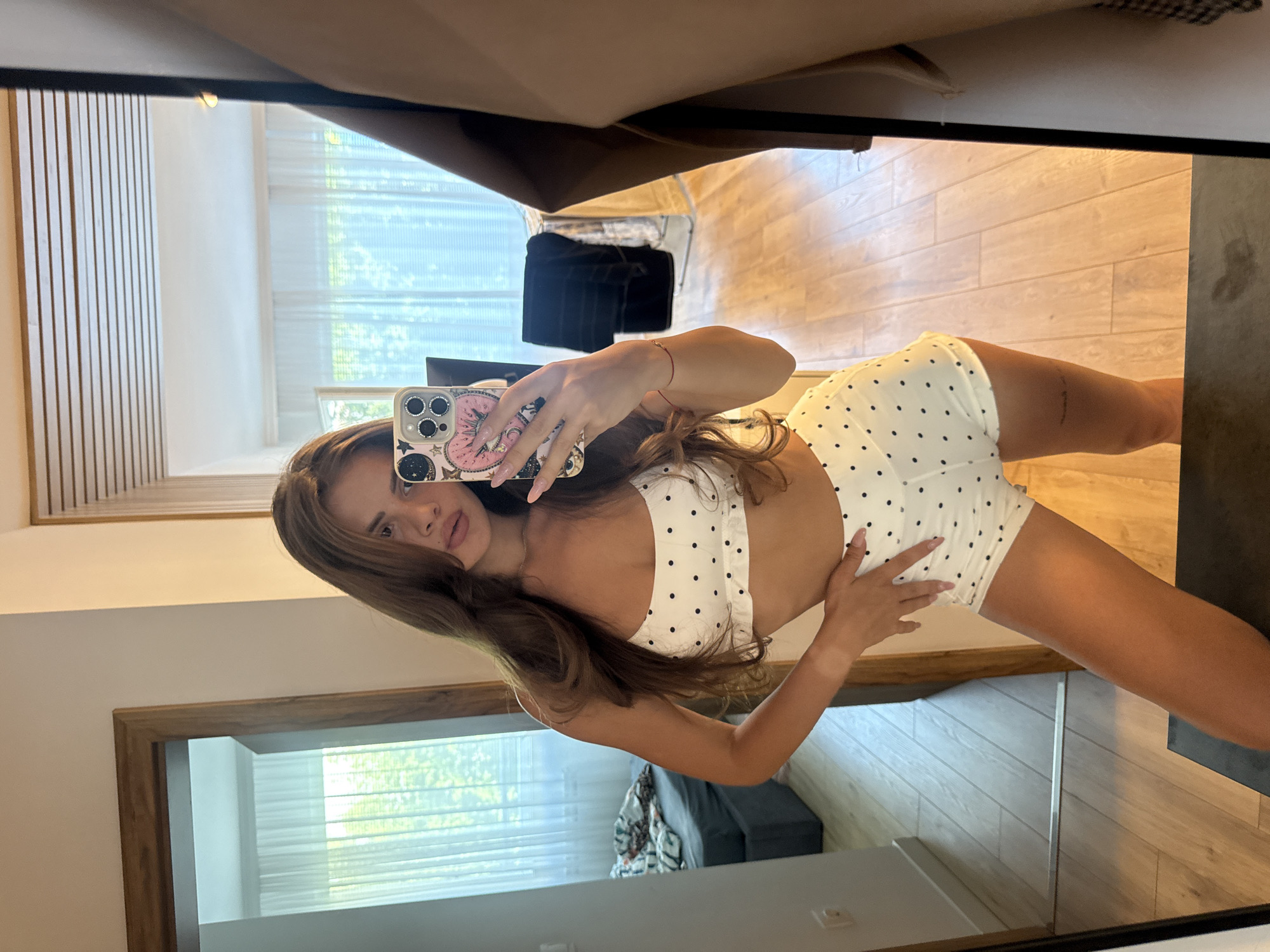A young woman with long brown hair is taking a mirror selfie. She is wearing a white polka dot bikini top and shorts. She is standing in what appears to be a bedroom, with a bed and a closet visible in the background. The room has wooden floors and white walls. The lighting is bright, likely from natural light coming through a window. Her phone is held up to take the picture, and she is looking at her reflection. The scene appears to be indoors, and there is no visible indication of the city of Plovdiv, Bulgaria.