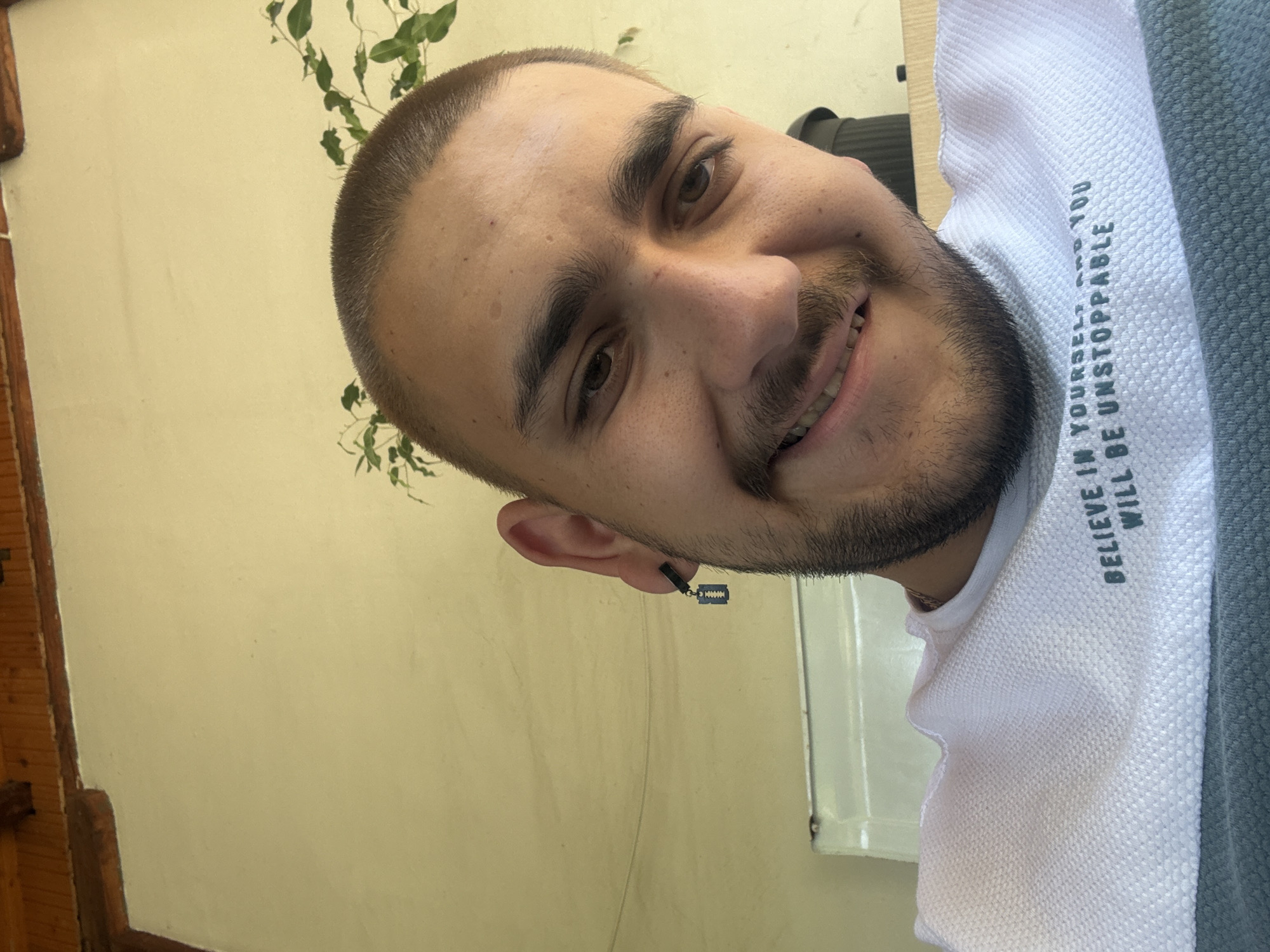 A close-up portrait of a young man, rotated 90 degrees clockwise, captures him smiling warmly at the camera. He has a short buzz cut, prominent dark eyebrows, a mustache, and a neatly trimmed beard. His white, textured t-shirt displays the motivational text, oriented vertically, which reads: "BELIEVE IN YOURSELF AND YOU WILL BE UNSTOPPABLE." Part of a grey garment is visible on his right side, possibly worn over the white shirt. A distinctive black earring with a miniature razor blade charm hangs from his left ear. The background features a plain, light yellow wall, with a wooden beam or frame visible on the far left. Green foliage, resembling a vine, is sparsely distributed in the upper left and behind his head. The natural lighting suggests a daytime setting. This image was captured in Staro Selo, Bulgaria.
