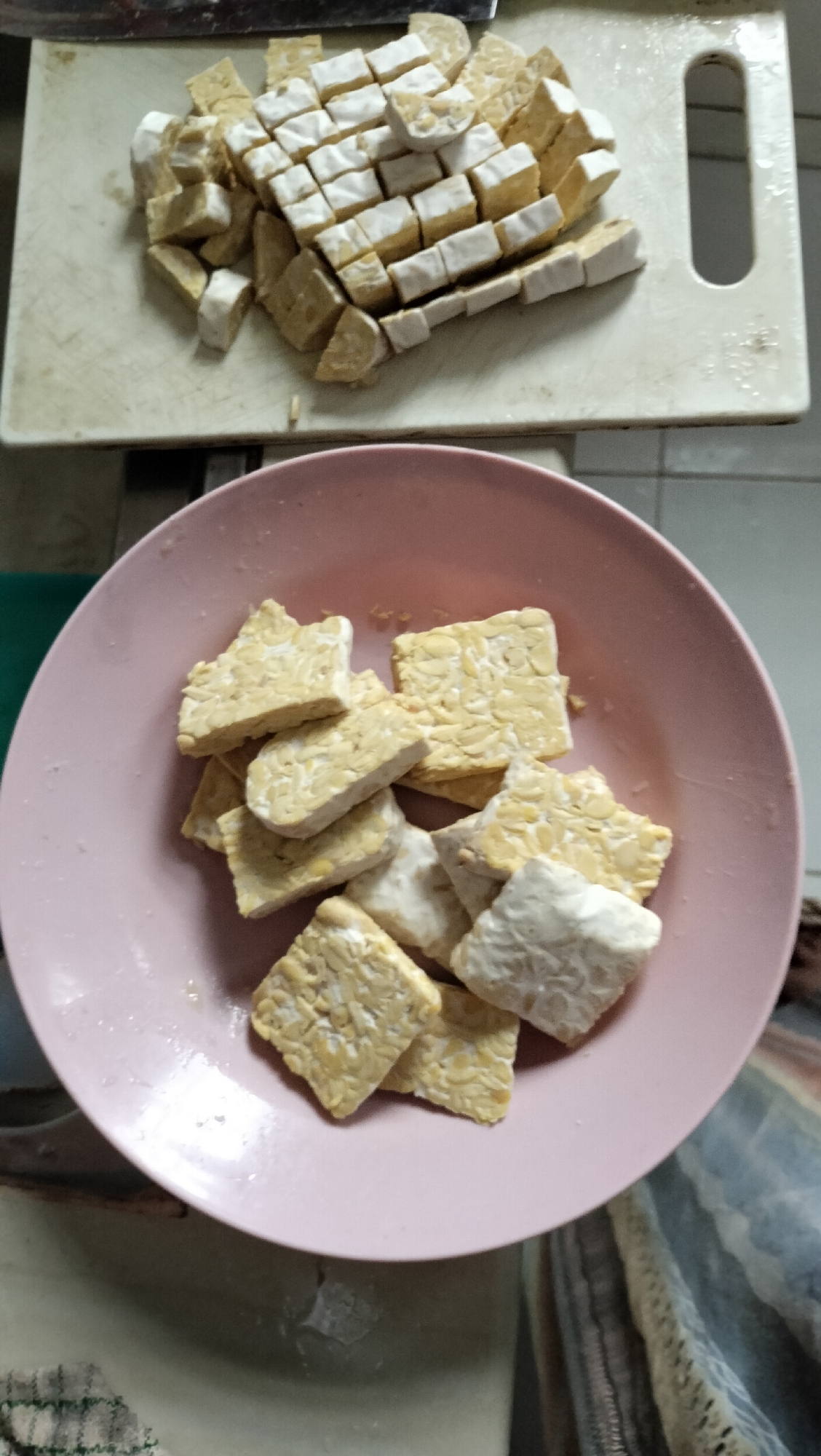 A close-up, top-down shot shows a pink bowl filled with cubed tempeh. The tempeh pieces are irregular in shape and size, some appearing lighter in color and more textured than others. They are arranged in a pile within the bowl, with some pieces overlapping. In the background, a white cutting board with more cubed tempeh is visible. The tempeh on the cutting board is more uniformly cut into small squares and rectangles. The overall scene suggests food preparation, likely in a kitchen. The image does not provide any specific clues to its location in Lhokseumawe, Indonesia.