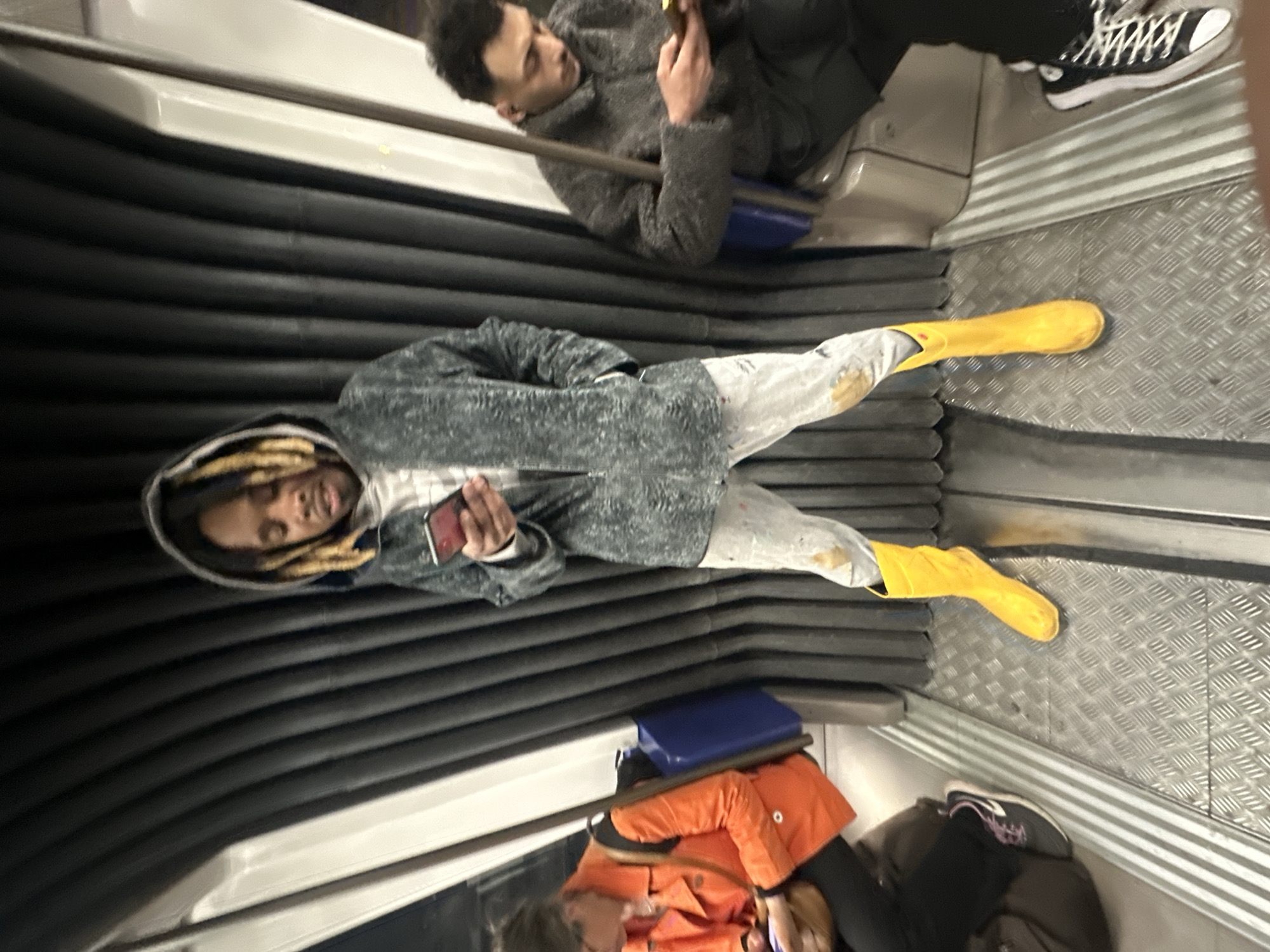 The image depicts the interior of a Paris Metro train. Three people are visible. One person, wearing a grey hooded jacket and bright yellow rubber boots, is sitting and looking at their phone. Another person is in a dark jacket and trousers and holding a phone. The third person, wearing an orange jacket, is also seated. The surroundings consist of the typical ribbed walls and metal flooring of the Metro carriage.