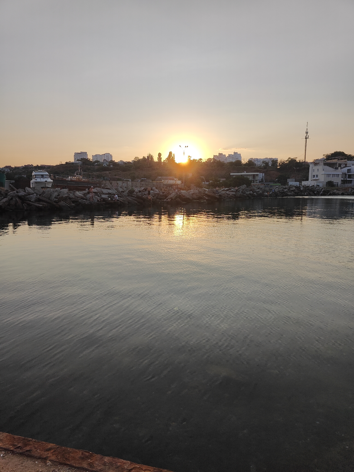The image shows a view of the Black Sea in Chornomorsk, Ukraine, during sunset. The sun is low on the horizon, casting a warm glow across the sky and reflecting on the water. In the foreground, the calm sea has gentle ripples. Along the shoreline, a rocky breakwater is visible, with several boats docked. Buildings and trees are situated on a hill in the background, overlooking the water. A few people can be seen near the boats on the breakwater. The overall scene depicts a peaceful evening at the harbor.