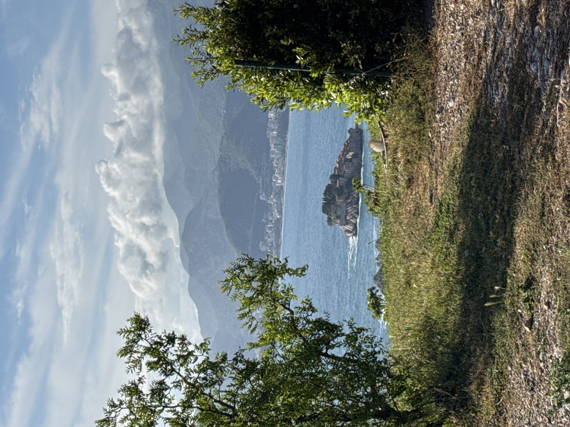 Captured from an elevated perspective in Blizikuće, Montenegro, the image showcases a coastal scene on a bright, sunny day. To the left, layered mountains fade into a blue sky streaked with white clouds. A coastal city spans along the shoreline. In the center of the image is a small island in the clear blue water. Green foliage frames the view in the foreground, both above and below the vantage point. A path of dirt and grass runs along the right side of the frame. No people are visible.