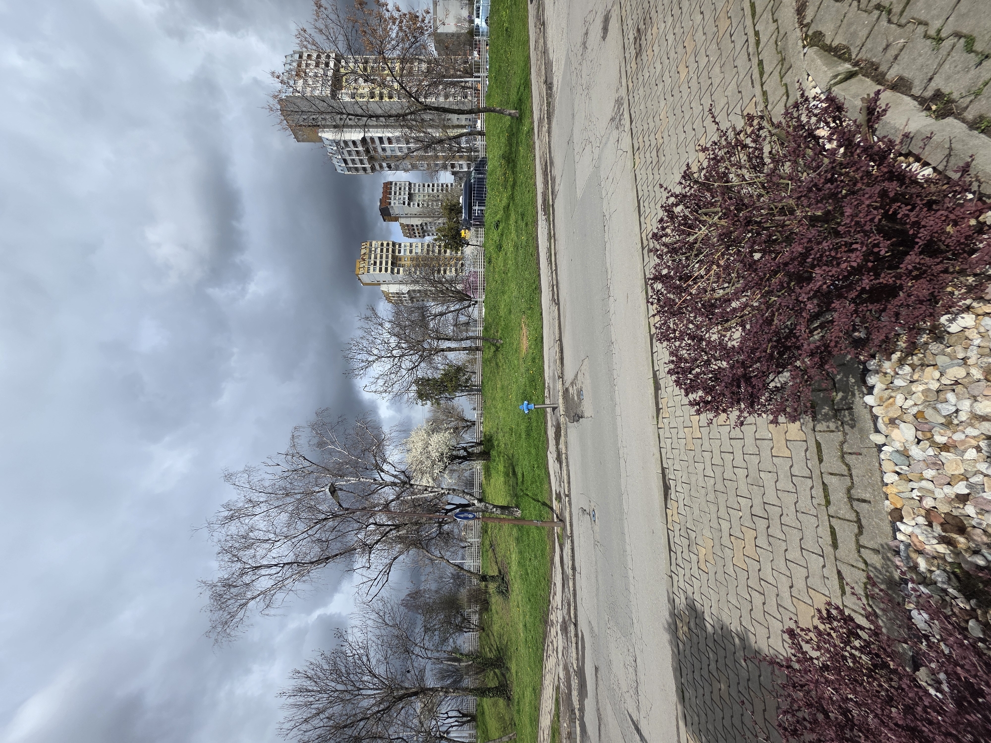 The image shows an urban landscape in Sofia, Bulgaria. High-rise residential buildings are visible in the background, under a cloudy sky. A green strip of grass separates the buildings from a pedestrian walkway constructed with interlocking paving stones. Bare trees and bushes are on the edges of the green area. The edge of the walkway is lined with pebbles. A blue fire hydrant is visible on the edge of the walkway. A blue and white sign is visible on a pole near the trees. The image appears to be taken from an elevated perspective.