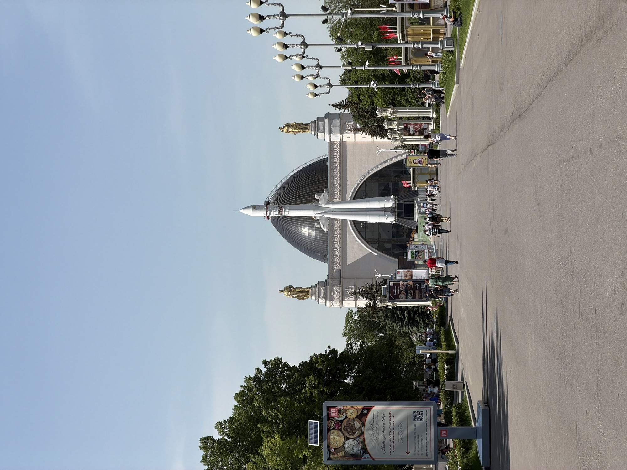 The image depicts the VDNKh (Exhibition of Achievements of National Economy) in Moscow, Russia, showcasing the iconic pavilion with a rocket on top. The rocket stands out against the backdrop of a clear blue sky. The pavilion itself is a grand structure with golden statues adorning its façade. In the foreground, there is a wide asphalt path with people walking along it. Along the side of the path, there are street lamps and advertising signs, one of which displays images of food, including what appear to be pastries, with Russian text and a QR code. Trees can be seen on the left side of the frame. Overall, the scene captures the bustling atmosphere of the VDNKh, a popular tourist destination in Moscow.