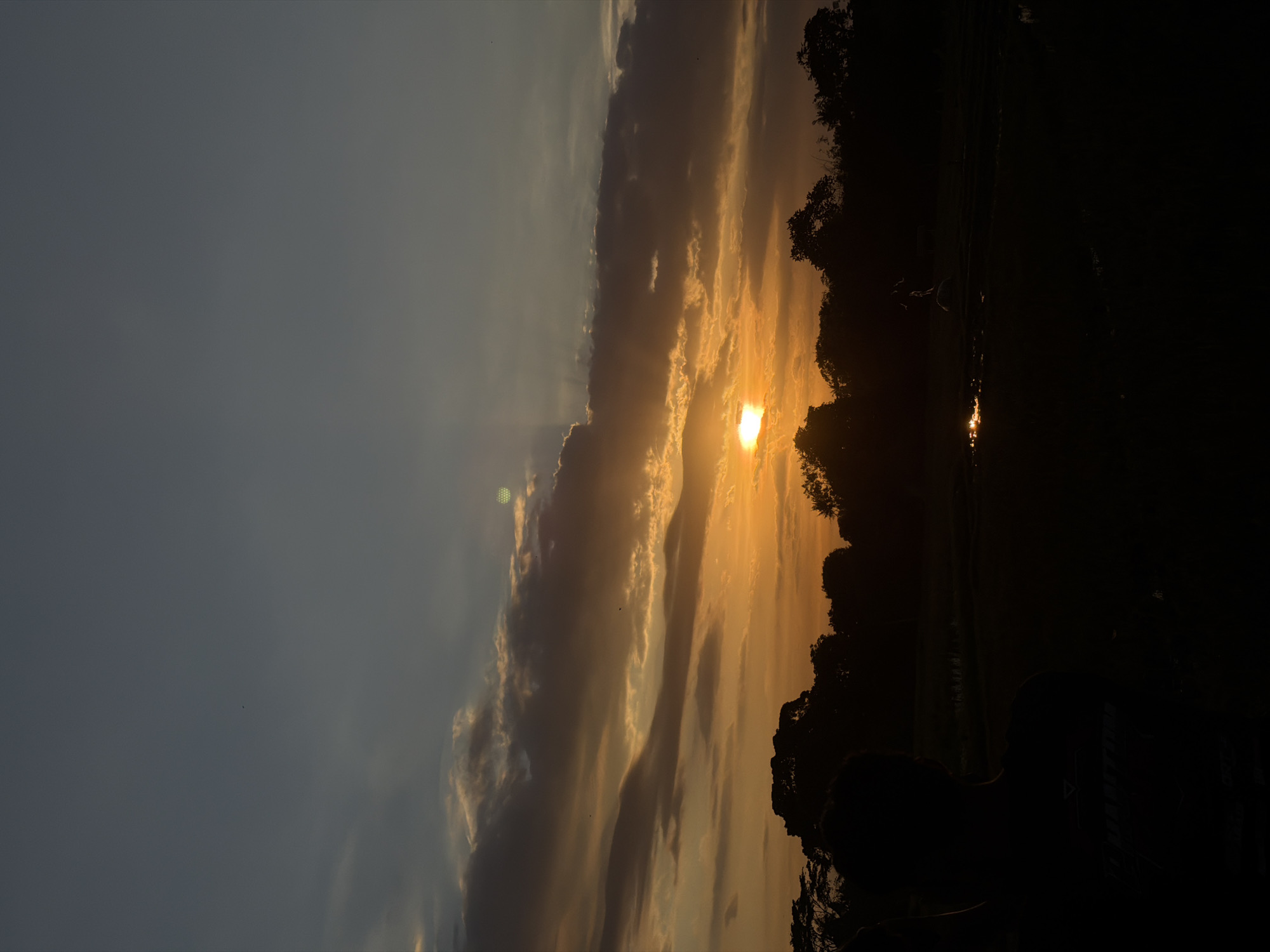 The image depicts a sunset scene, possibly in North Kuta, Indonesia. The sun is obscured by clouds, casting a golden glow on the sky and reflecting off what appears to be water. The silhouetted trees and other dark objects are on the right side of the image with a person in the foreground. The sky transitions from a blue hue on the left to an orange hue as it gets closer to the sun. The photo is taken at a rotated angle.