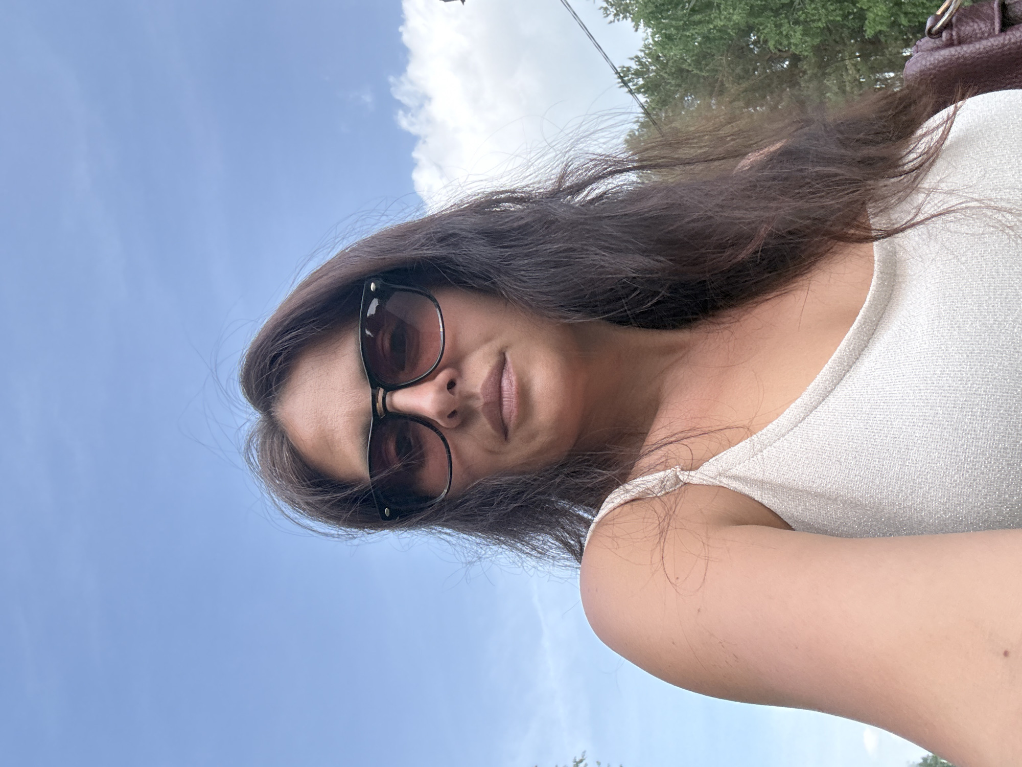 A woman with long brown hair wearing sunglasses and a white tank top is photographed from a low angle against a background of blue sky with some clouds and a partially visible tree. A brown handbag is visible in the upper right corner of the frame. The location is unknown and cannot be determined from this image.