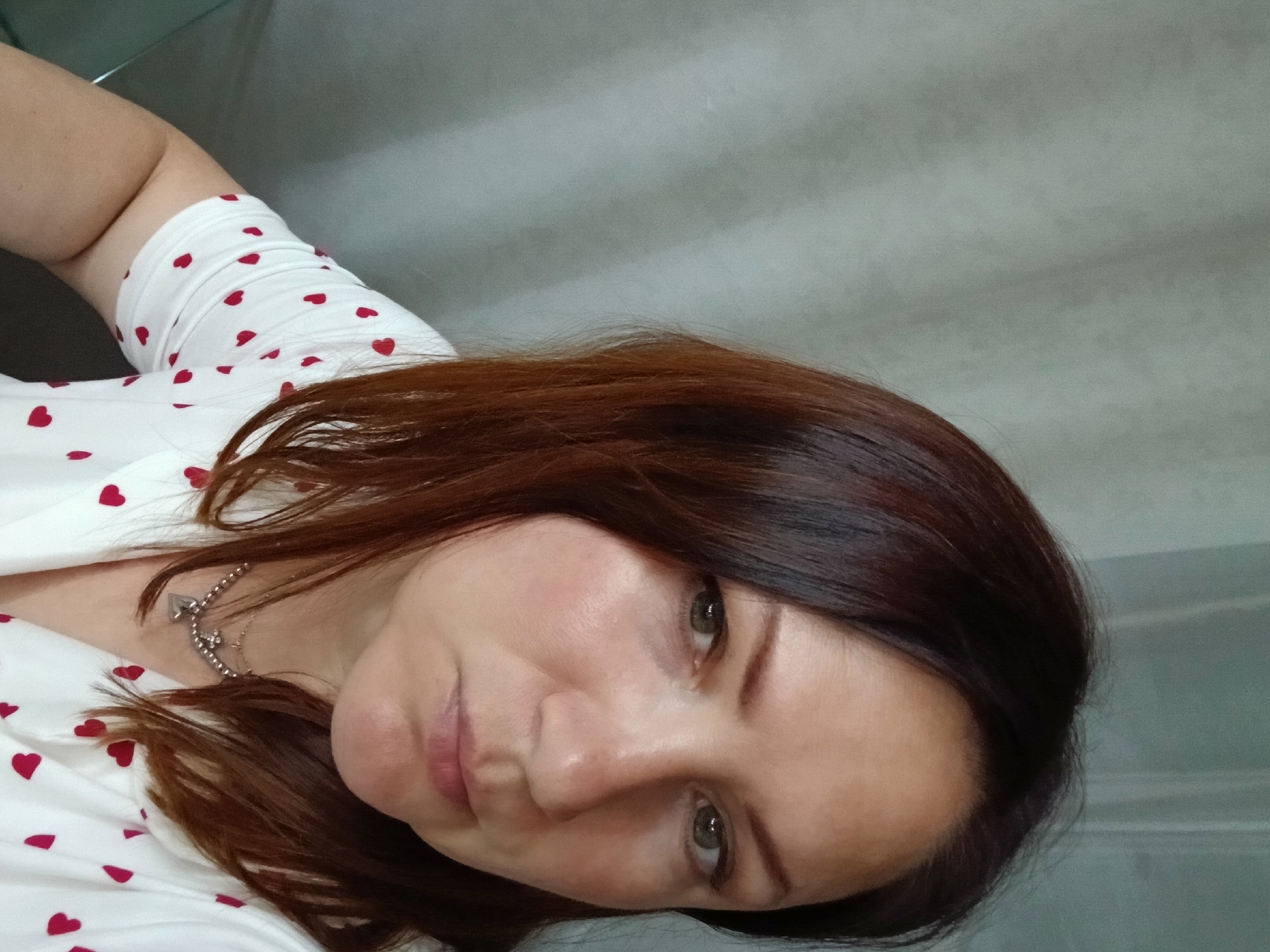 A medium shot shows a fair-skinned woman with auburn hair and green eyes. She is wearing a white shirt with small red hearts and a silver necklace. The background is a pale gray wall. The image appears to be a selfie. The woman is looking towards the camera, with a neutral expression. Her head is tilted slightly to her left. She has a light dusting of blush on her cheeks and her lips are slightly parted. The lighting is soft and diffused, suggesting it is daytime. The overall scene is domestic and casual. There are no visible landmarks or clues to indicate the specific location in Chaikovsky, Russia.