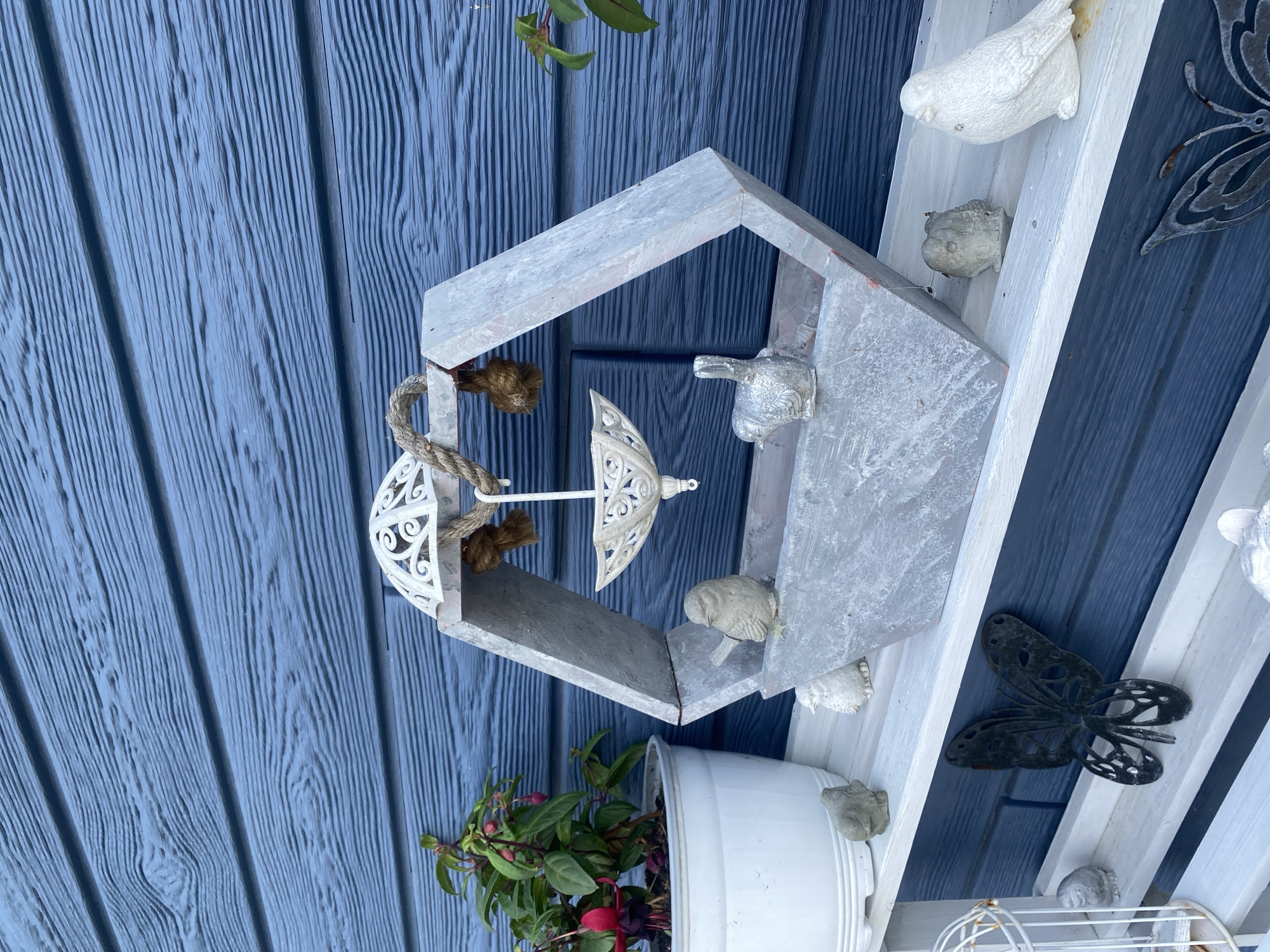 The image depicts an outdoor scene against a blue, wood-paneled wall. On a white shelf, there is a hexagonal structure made of gray, weathered material, possibly concrete or stone. This structure has an open top and a smaller decorative element suspended from it by rope. Several small bird figurines are placed around and on the hexagonal structure. To the left of the structure is a white pot containing a flowering plant with pink and red blossoms. Further to the left, a white birdcage is partially visible. On the shelf below, a black metal butterfly decoration is prominent. The overall impression is one of a decorative garden display. There is no discernible indication of the location being in Tret'ya Pad', Russia.