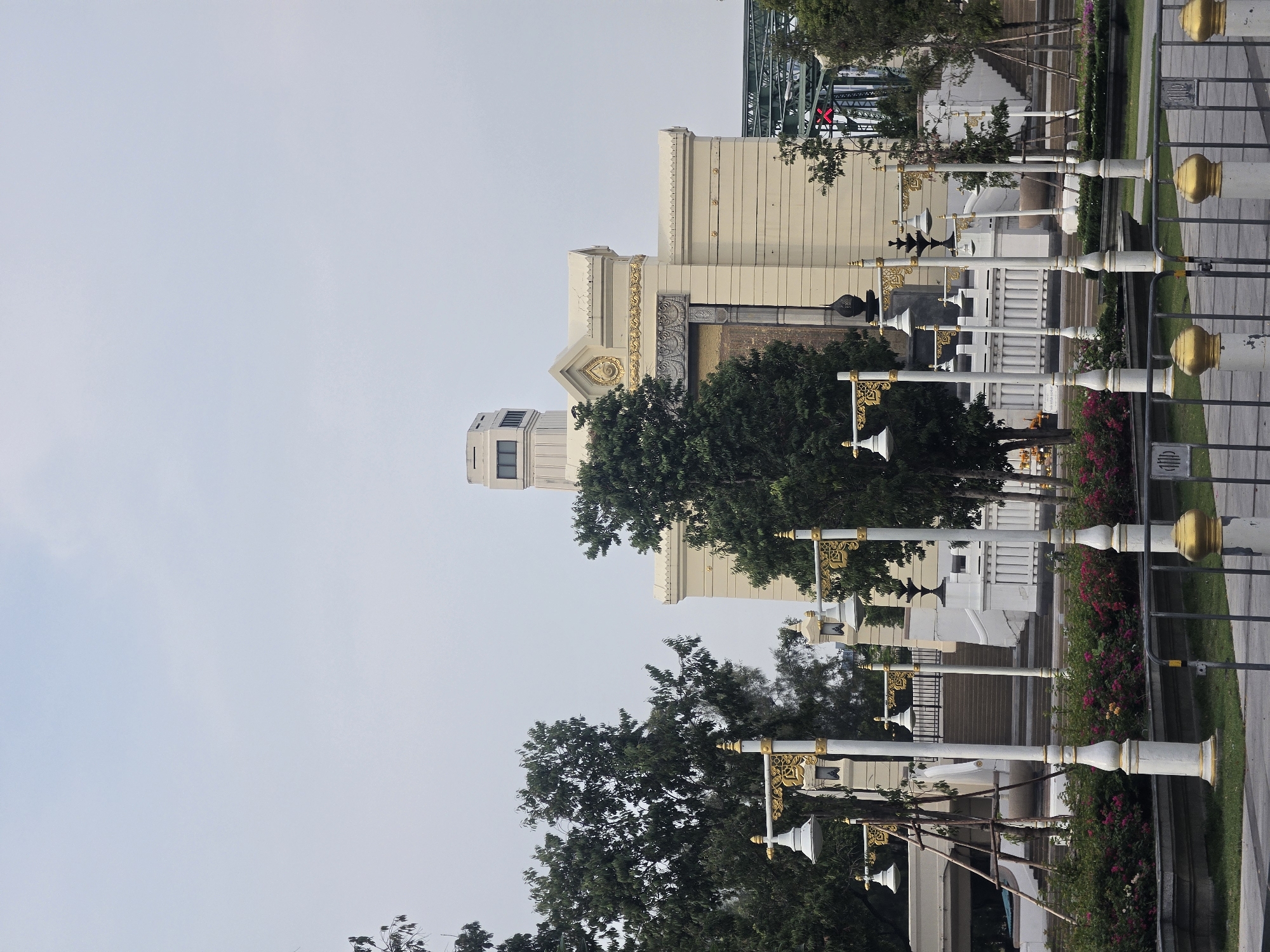 The image captures a view of Phra Sumen Fort in Bangkok, Thailand. The fort, a beige, fortress-like structure with a distinct tower and ornate details, is partially obscured by green trees. The fort stands behind a row of white, decorative lampposts, some with gold accents. The ground level features vibrant red and pink flowering bushes. The Chao Phraya River's bridge is visible in the background. The sky is a pale grey color.