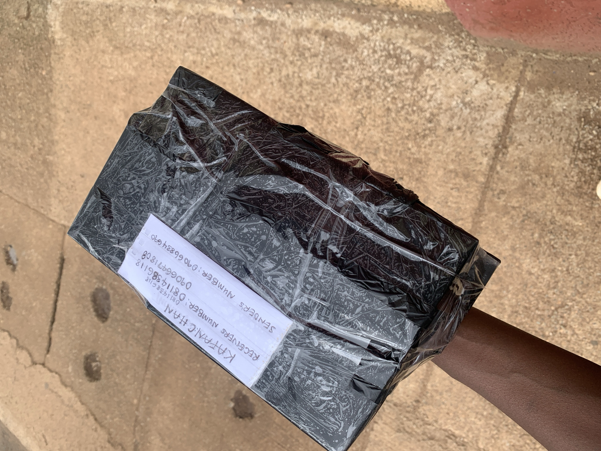 A package wrapped in black plastic is held by a person's arm and hand, against a background of cracked concrete paving stones. A white label is attached to the package, with handwritten text that reads "KAFA.N.CH.AN", "RECEIVER'S NUMBER: 08114386118", and "SENDER'S NUMBER: 09066834690". The image was taken outdoors in the city of Jos, Nigeria.
