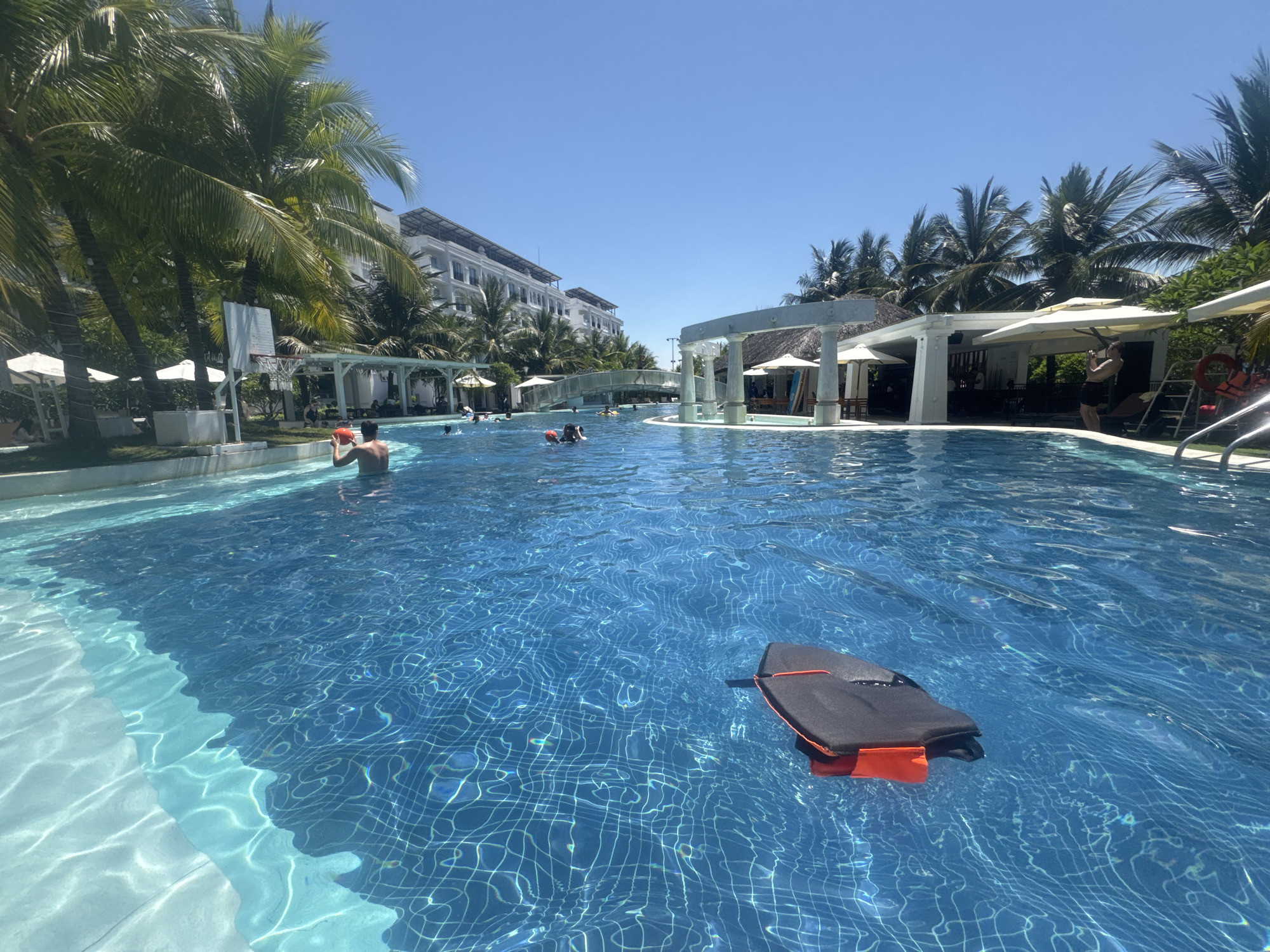 This image depicts a swimming pool area with a view of a multi-story white building and lush palm trees, indicative of a tropical resort. The pool is a vibrant blue, with sunlight creating shimmering patterns on the tiled bottom. Several people are visible in the water, some swimming and others standing. In the foreground, a black and orange flotation device is partially submerged. In the background, there is a covered seating area with umbrellas and a white bridge spanning across a section of the pool. A basketball hoop is also visible near the edge of the pool. The sky is clear and blue. This scene is likely from a resort in Bac Nha Trang Ward, Vietnam, known for its beautiful beaches and resorts.