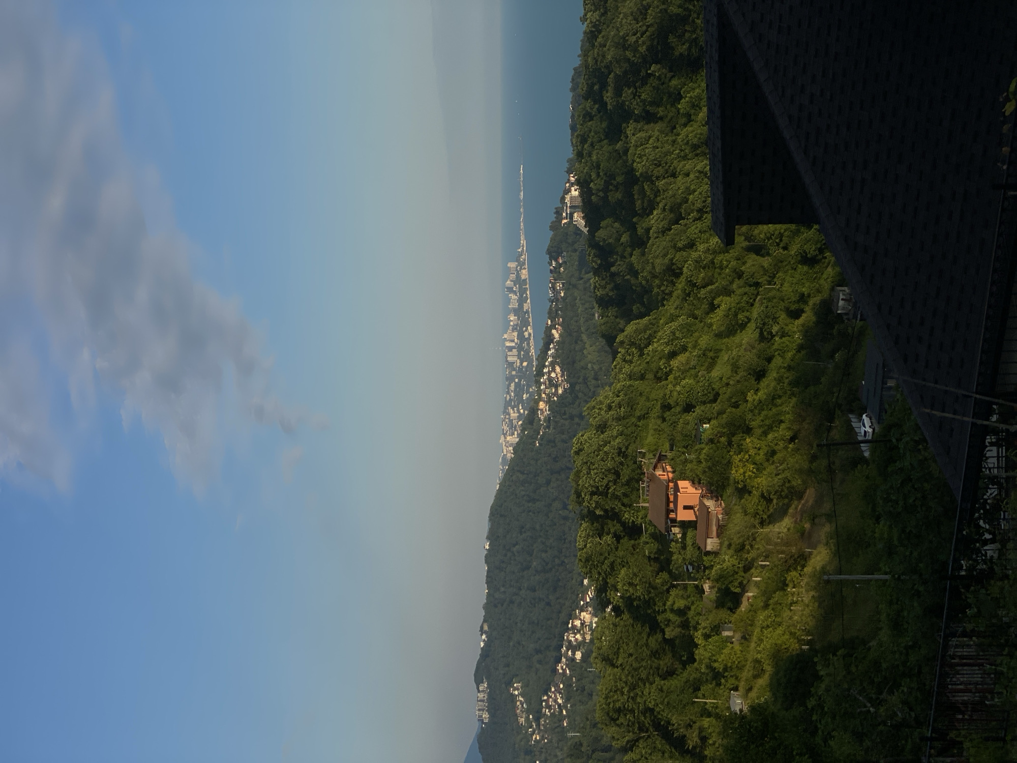 The image shows a panoramic view of Dagomys, Russia, overlooking the Black Sea. In the foreground, the dark, tiled roof of a building dominates the bottom right corner. Beyond that, a hillside covered in lush green trees slopes downwards. A bright orange house with a red roof is nestled on this hillside, surrounded by more dense vegetation.
Further in the distance, the green slopes continue, dotted with clusters of white and light-colored buildings, suggesting a residential area. The middle ground is filled with rolling green hills, and the distant background features a coastal city with numerous tall buildings stretching along the shoreline. The Black Sea occupies the upper right portion of the image, its surface calm and reflecting the pale blue sky. A few faint clouds are visible in the sky. The overall scene depicts a serene, sunlit day in a coastal mountainous region.