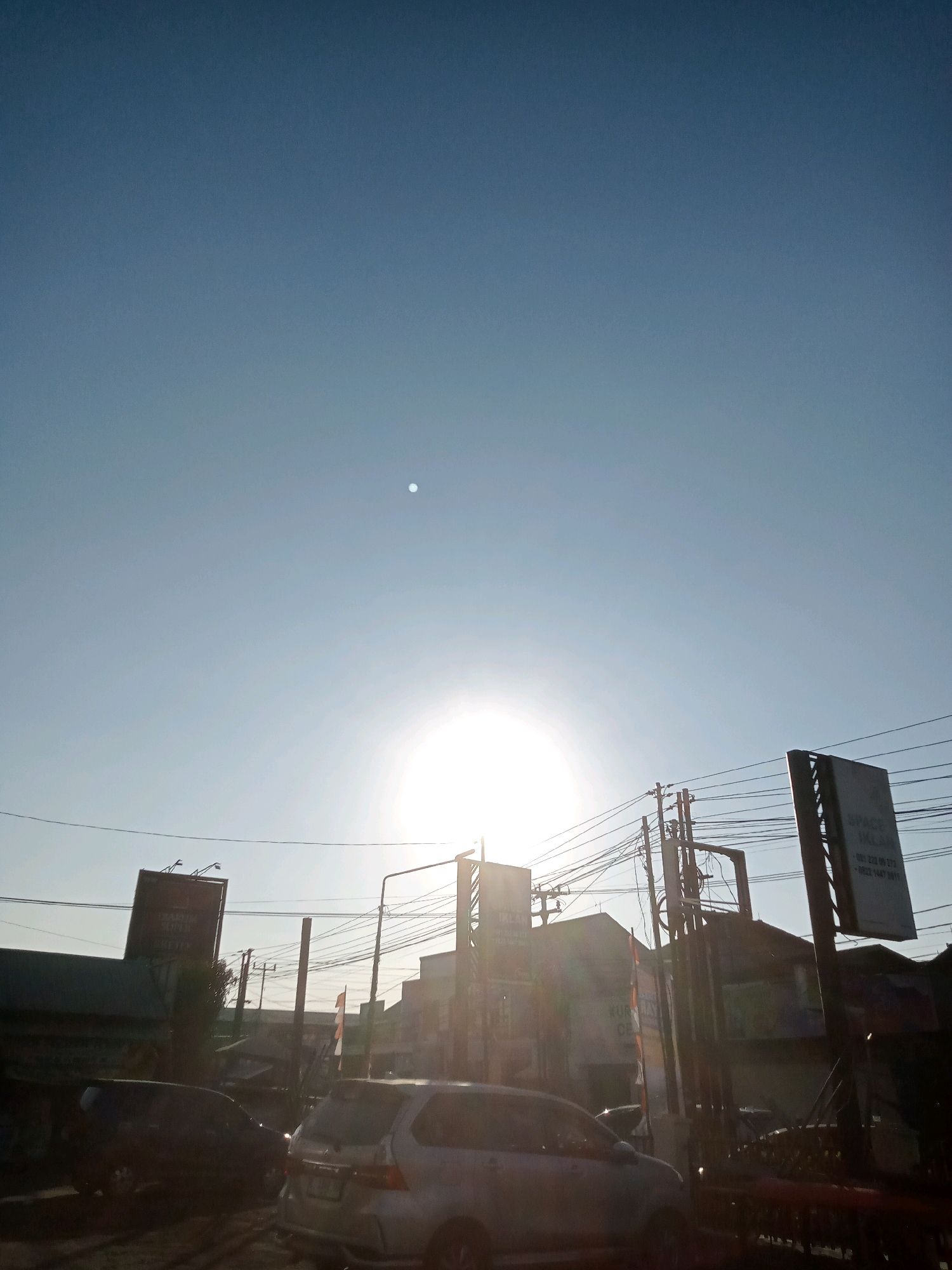 The image depicts a street scene in Cirebon, Indonesia, during what appears to be sunrise or sunset, given the low angle of the sun and the clear sky. The sun is a bright, hazy orb positioned in the upper middle portion of the frame, casting a strong glare. Several utility poles with a dense network of wires are visible across the mid-ground, suggesting an urban or semi-urban environment.
Buildings are present on either side of the street, with some appearing to be commercial establishments indicated by signage, though much of it is illegible due to the glare and distance. One prominent sign on the right reads "SPACE IKLAN" which translates to "Advertising Space." Vehicles are present on the road, including a silver minivan in the foreground on the right and a dark-colored SUV or van on the left. The vehicles are stopped or moving slowly, suggesting traffic.
The overall impression is a typical street view in a populated area, characterized by infrastructure, vehicles, and signs, under the bright illumination of the sun. The sky is a gradient of pale blue, becoming darker towards the top.