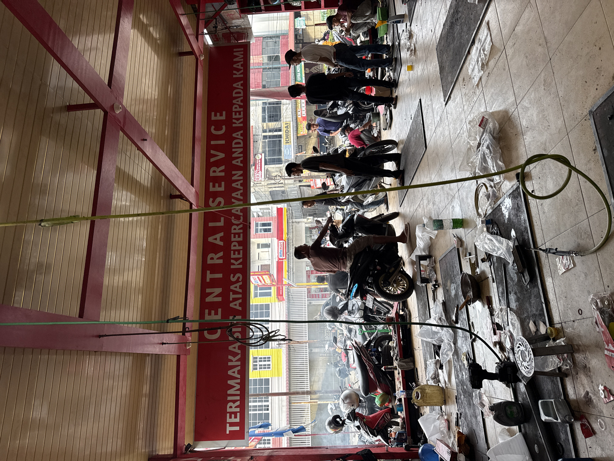 The image shows the interior of a motorcycle repair shop. Several men are working on various motorcycles. The floor is tiled. Tools and motorcycle parts are scattered on the floor. A sign on the wall reads "CENTRAL SERVICE TERIMAKASIH ATAS KEPERCAYAAN ANDA KEPADA KAMI" (Thank you for your trust). Outside the shop, buildings are visible, one with an Alfamart sign, indicating the location as Lhokseumawe, Indonesia.