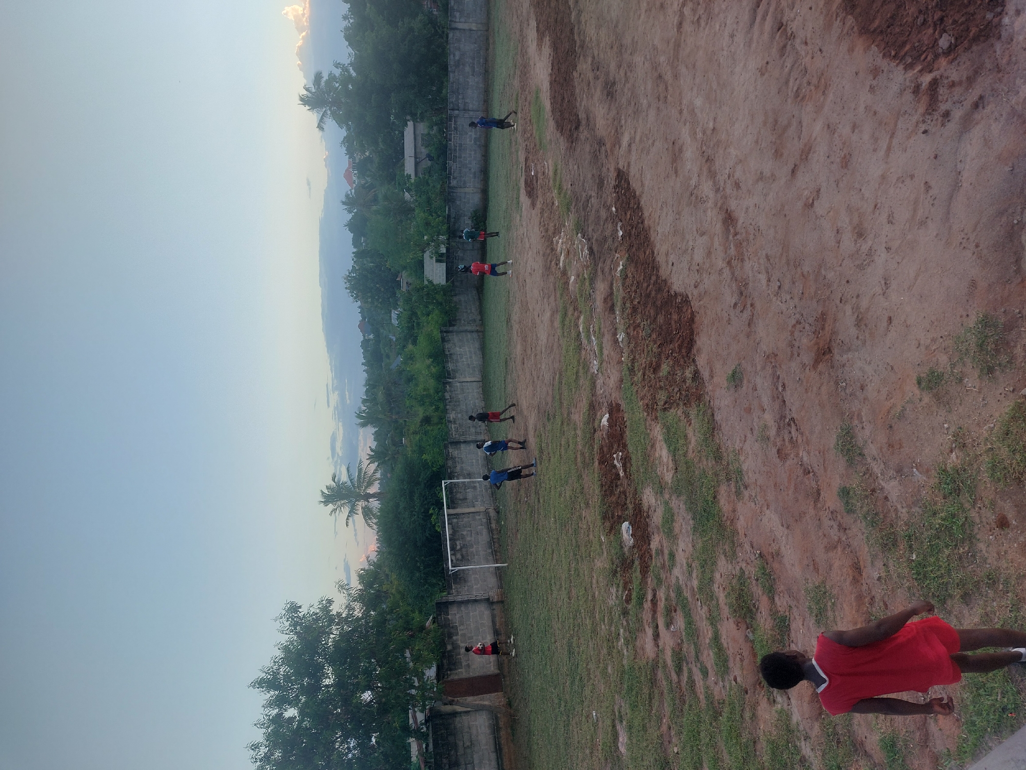 A medium shot reveals a naturalistic outdoor soccer field in Dar es Salaam, Tanzania. The foreground features a dry, reddish-brown dirt surface with scattered patches of green grass. A young man wearing a red shirt and dark shorts is walking in the lower right corner of the frame, his back partially visible to the viewer. Further into the scene, several individuals are seen practicing or playing soccer on a stretch of green grass along the field's edge. A standard white soccer goal is centered along the tree and foliage lined edge. A tall concrete fence is located behind this. In the background, a blend of lush trees, vegetation, and distant residential buildings can be observed, which are overshadowed by a gradient of sky transitioning from light blue near the horizon to a paler hue at the top of the frame. The lighting suggests either early morning or late afternoon.