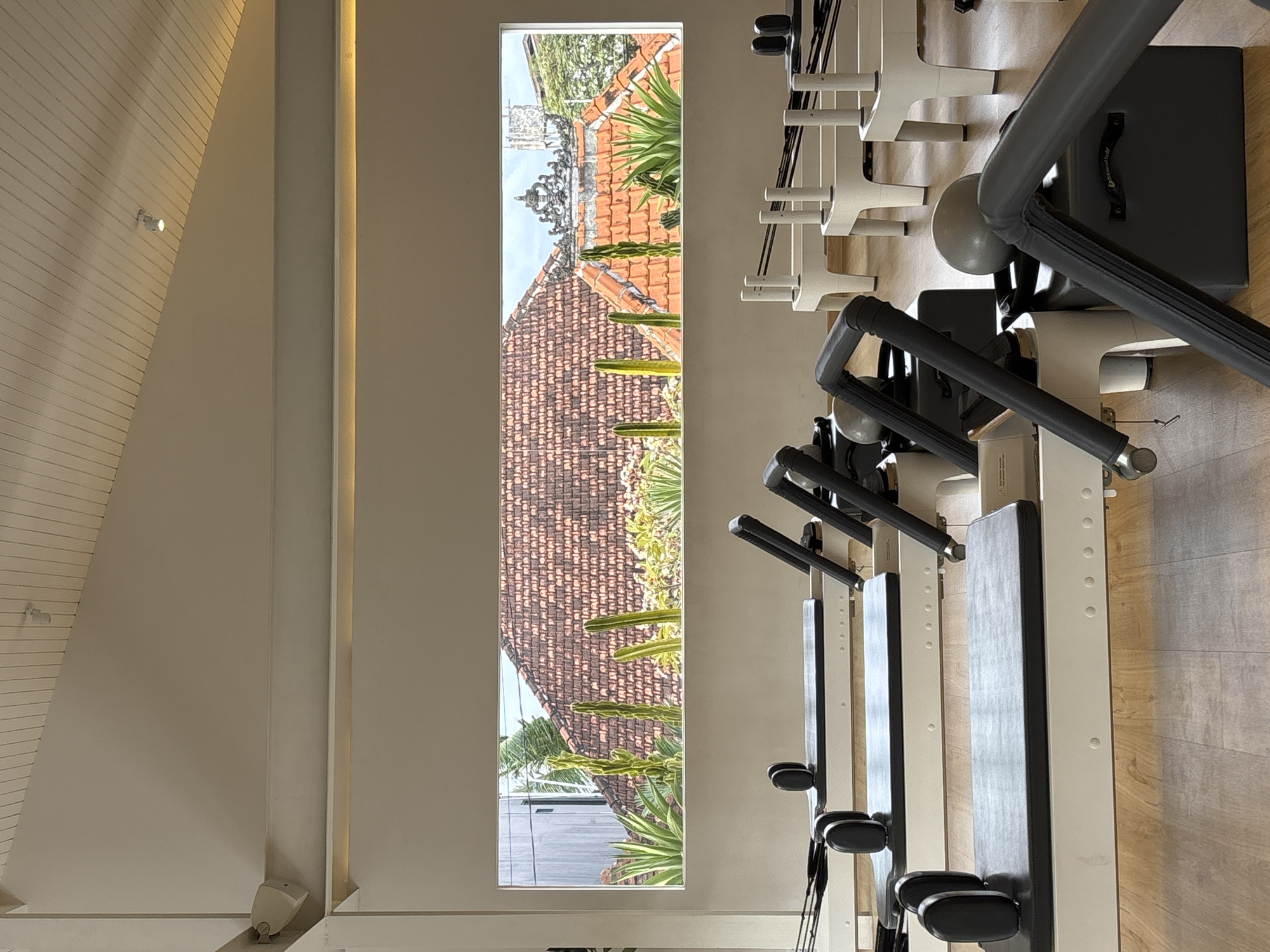 The image shows an interior view of a building, possibly a Pilates studio, in North Kuta, Indonesia. The prominent features are several Pilates reformer machines lined up, viewed at an angle. A tall, narrow window offers a glimpse outside, revealing a landscape of traditional Indonesian architecture with orange-tiled roofs and tropical greenery. The interior walls are light-colored, with subtle design elements such as vertical strips and possibly wood-paneled ceilings. The lighting appears to be soft and natural. The floor is wooden. There are no people visible.
