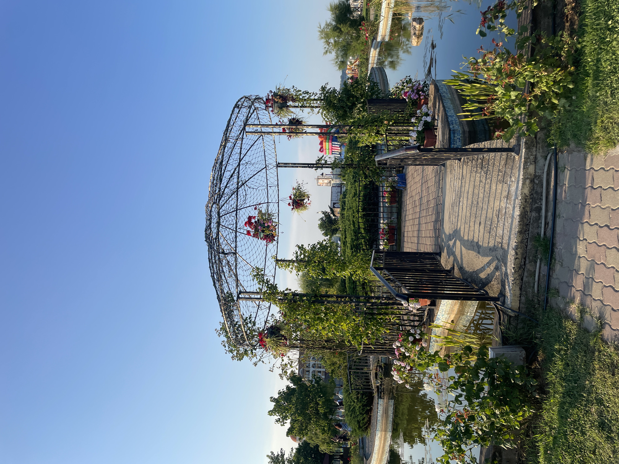 The image shows a decorative gazebo in a park-like setting, likely located in the city of Obzor, Bulgaria. The gazebo is an open, circular structure with an intricate metalwork frame, adorned with climbing vines and hanging flower baskets filled with red and pink blooms. A small arched bridge leads to the gazebo, which is situated on a small island in a calm body of water, possibly a pond or canal. The water reflects the surrounding greenery and the clear blue sky.
Along the water's edge and the bridge, there are numerous potted flowers, primarily white and pink roses, and tall green reeds. A black metal railing lines the bridge and the perimeter of the gazebo's island. In the background, there are trees, bushes, and buildings, suggesting an urban or resort area. A red-domed building is visible in the distance, a common architectural feature in many Bulgarian coastal towns. Further back, a brightly colored inflatable structure, resembling a bouncy castle, is partially visible, indicating a recreational area. The foreground shows a paved walkway made of interlocking concrete pavers, with grass on either side. The lighting suggests it is late afternoon or early morning, with long shadows cast by the gazebo and railing onto the walkway. There are a few people visible in the mid-ground, casually strolling through the park, but they are not the primary focus of the image. The overall scene conveys a sense of peaceful outdoor leisure.