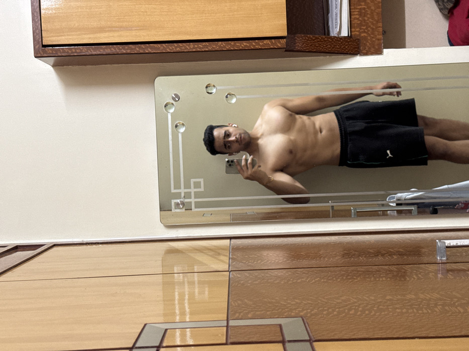 The image is a mirror selfie taken indoors, rotated 90 degrees clockwise. When viewed upright, it shows a shirtless man reflected in a decorative rectangular mirror.
The man, appearing to be of a youthful to middle age, has dark hair neatly styled and a clean-shaven or lightly stubbled face. He is wearing small, white wireless earbuds in both ears. He displays a toned physique, showing his chest, abs, and arms. He is wearing black athletic shorts with a white logo, which resembles the PUMA brand, visible on the left thigh area of the shorts. On his left wrist (right in the reflection), he wears a thin, gold-colored bracelet or watch. He is holding a modern smartphone with a light-colored case (possibly white or light grey) in his right hand (left in the reflection), using it to capture the selfie. The phone's multiple camera lenses are visible on its back. The man is looking directly at his reflection in the phone, with a neutral to slightly serious expression.
The mirror itself has a decorative etched design along its borders, featuring geometric patterns such as straight lines and squares, particularly prominent in the corners. Several round, clear, raised embellishments, possibly glass studs or cabochons, are affixed to the mirror's surface in the corners and along the top edge of the decorative border.
Outside the mirror's reflection, the background shows parts of a room. Above and to the left of the mirror is a plain, light-colored wall (off-white or cream). Directly above the mirror, a wooden cabinet or shelf with a warm, reddish-brown hue and visible wood grain is mounted. Items, possibly papers or books, are partially visible inside the right section of this cabinet. Below the mirror, there's a polished wooden surface, which appears to be a floor or a large piece of furniture like a dresser. This surface features a glossy finish and a geometric pattern created by lighter and darker wood sections, forming a tiled or paneled effect, including a distinct square-within-a-square design. A faint reflection of the room is visible on this glossy wooden surface. In the bottom right corner of the mirror's reflection, some folded white and blue fabric, possibly laundry or a towel, is visible.
The lighting in the room appears bright and even, likely from an overhead source or an unseen window, creating a well-lit environment for the selfie. The overall setting suggests a private, residential space.