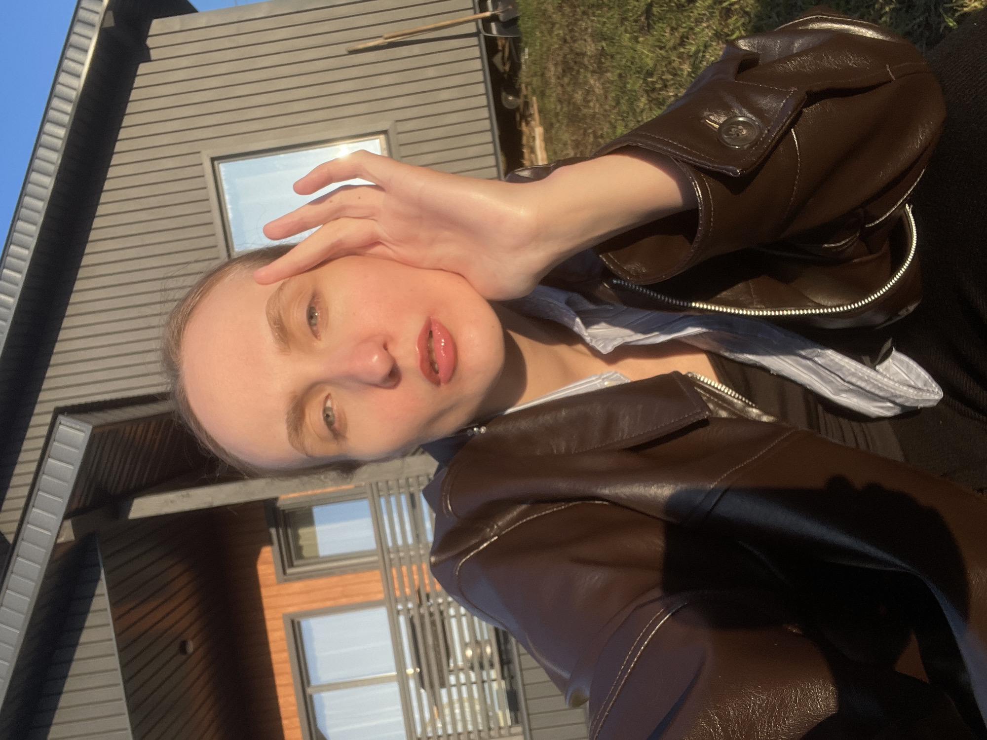 A rotated selfie-style image shows a person outdoors on a sunny day. The image is oriented with the person's head to the right, suggesting it was taken at a Dutch angle and rotated clockwise for viewing. The subject is a person, likely female, with fair skin, light eyebrows, and partially visible eyes. Their right hand is raised, with fingers resting near their forehead/hairline. They have slightly parted lips.
The person is wearing a dark brown, possibly leather or faux leather, zippered jacket over a light-colored striped shirt. Strong natural light illuminates their face and jacket, creating reflections and highlighting details like the metal zipper and a decorative button on the jacket's lapel.
In the background is a modern-style building with dark gray horizontal siding and several rectangular windows, which reflect the bright blue sky. Green grass or vegetation is visible to the upper right of the person, completing the outdoor setting. The bright sunlight and clear sky indicate daytime with good weather. No text is visible in the image. The location context is Sovetsko-Nikol'skoe Lesnichestvo, Russia.