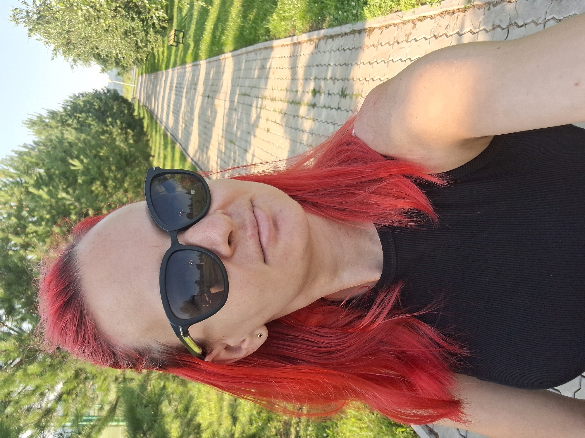 A medium shot shows a woman with bright red hair, wearing black sunglasses and a black ribbed tank top. She is outdoors, standing on a paved walkway with a lawn and trees on either side. In the distance, the walkway stretches out lined with trees, and there is a bench visible on the right side of the walkway. The sun is shining, casting shadows on the walkway and illuminating the woman's arm with a warm glow. The background suggests a park or recreational area. The reflection in her sunglasses shows a clear sky and possibly trees. The location is identified as Atamanovka, Russia.