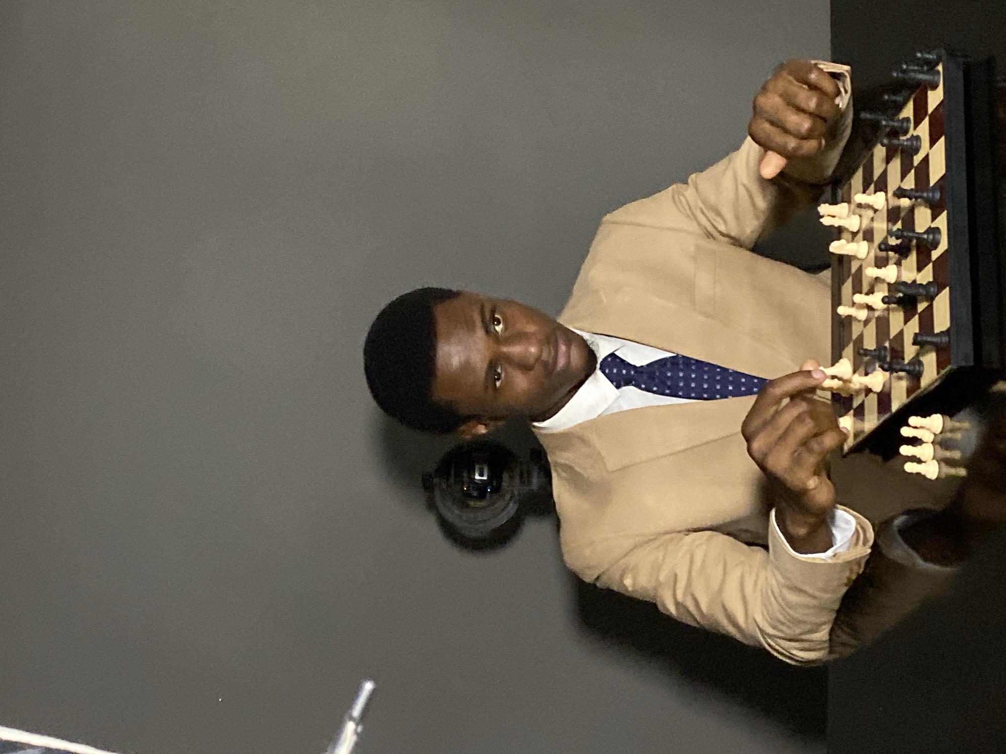 A man is posing with a chess set. He is wearing a tan suit with a white shirt and blue tie. He is holding the chess pieces. The background is a gray wall. The location is Gura Top, Nigeria.
