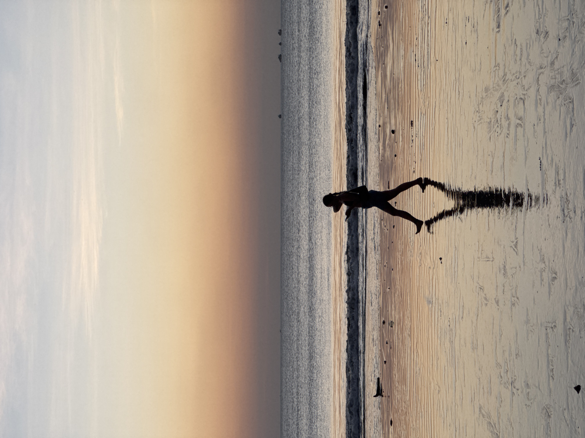 The image shows a person walking on a beach in Kuta Selatan, Indonesia, during sunset. The sky is a gradient of soft orange and pale blue, with wispy clouds. The ocean is calm, with gentle waves breaking on the shore. The wet sand of the beach reflects the colors of the sky and sea, creating a shimmering effect. In the distance, several boats are visible on the horizon. The person is silhouetted against the sunset, walking from left to right. They appear to be wearing shorts and a tank top. Their reflection is clearly visible in the wet sand. The overall atmosphere of the image is peaceful and serene.