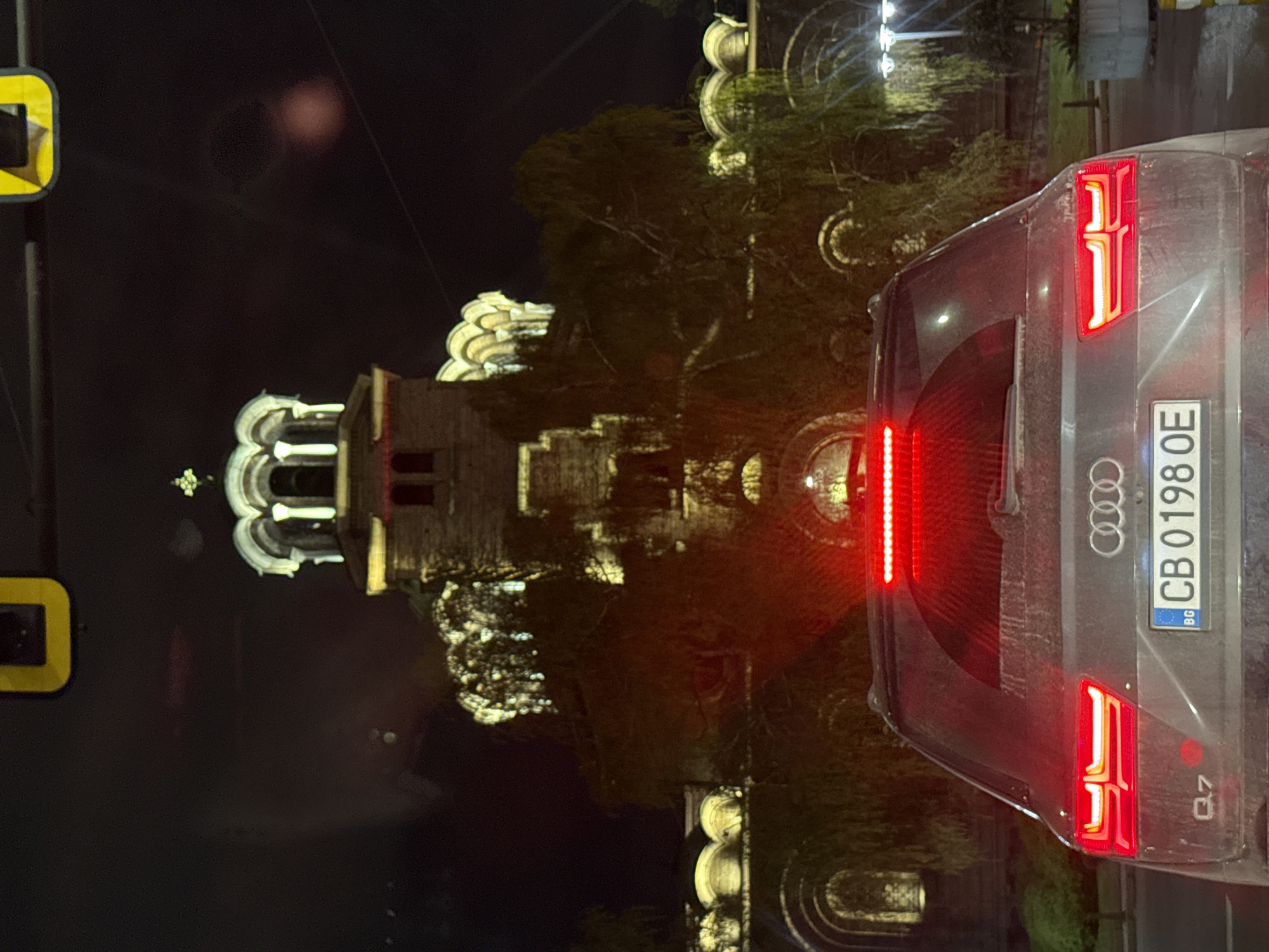At night, a gray Audi Q7 with Bulgarian license plate "CB 0198 OE" is parked on a street. Illuminated buildings and trees are behind the car. A yellow sign is visible to the left. The location is in Unknown, Unknown.