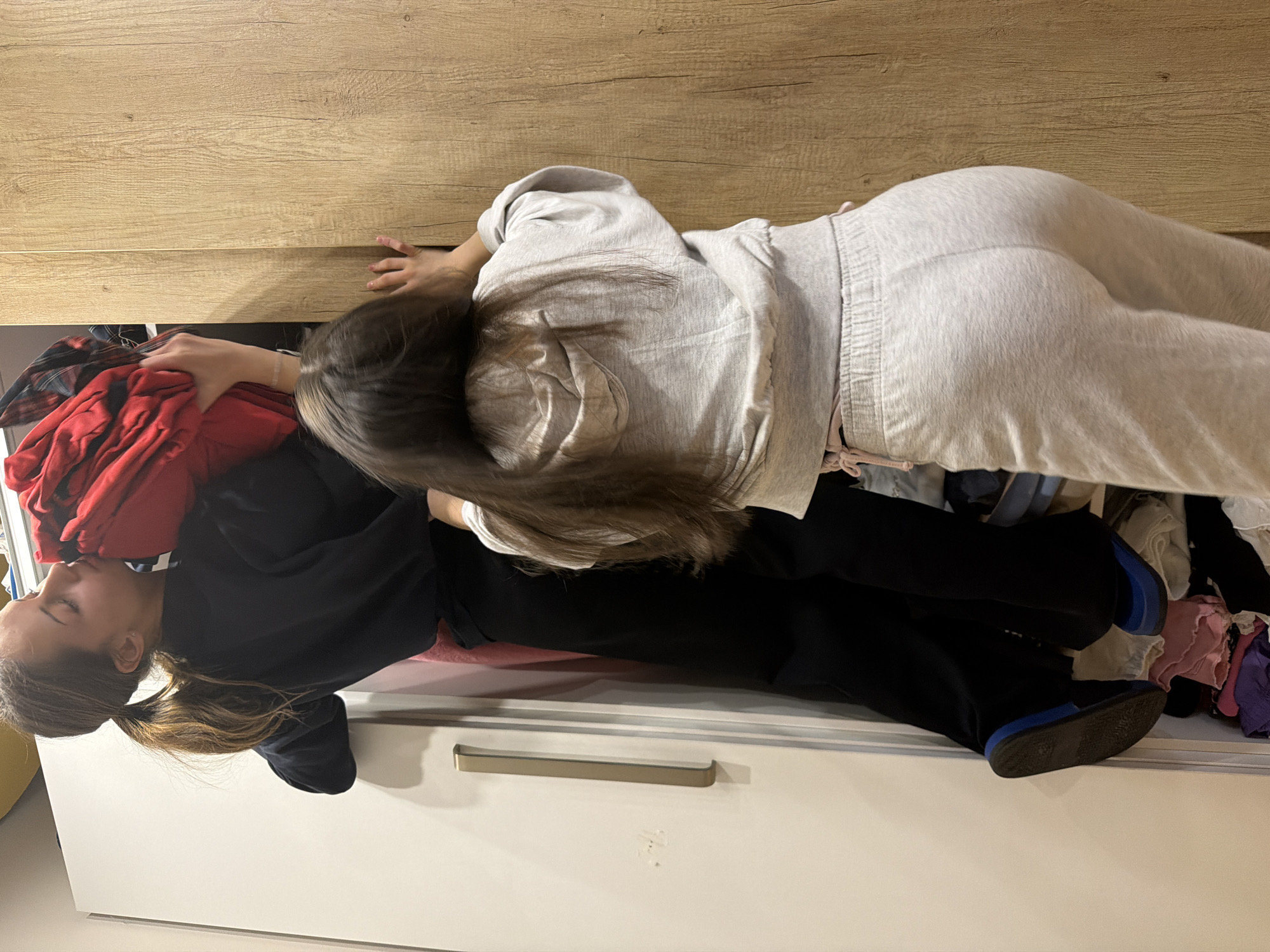 This indoor scene, located in Sofia, Bulgaria, depicts two young females in proximity to a wardrobe system. The individual on the left, wearing a dark long-sleeved top, dark pants, and blue sandals, is positioned with her left foot inside an open white wardrobe compartment containing stacked clothing. Her right arm is elevated, holding a bunched red garment near the top of the wardrobe, with her head tilted, resting her face against the fabric. Her hair is styled in a ponytail. The second individual, positioned on the right and viewed from behind, wears a light grey hooded sweatshirt and matching sweatpants. Her hands are placed on the light wood-paneled surface of an adjacent wardrobe door. The wardrobe system comprises a white unit on the left, featuring a vertical gold-colored handle, and a light wood-paneled unit on the right.