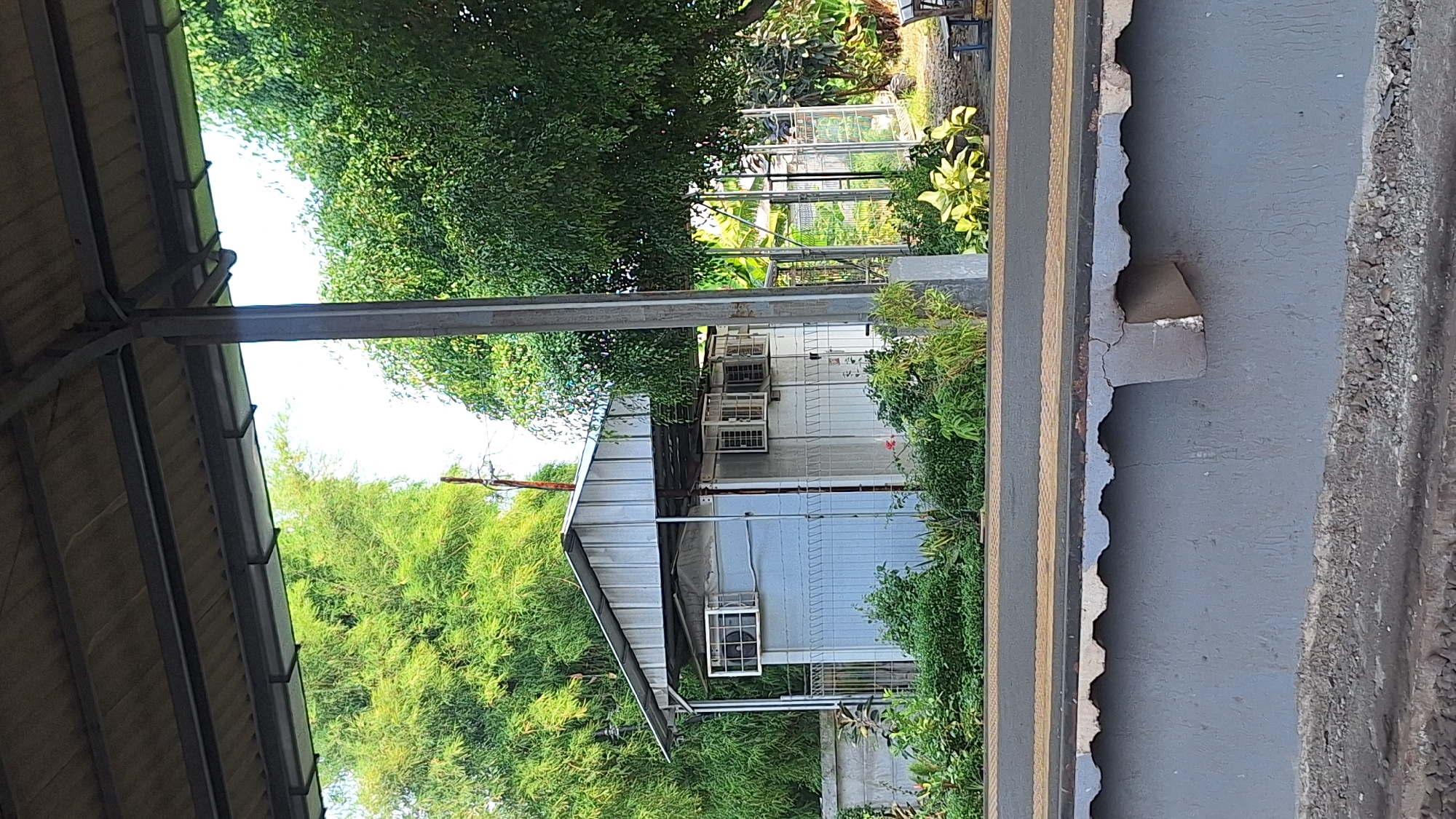 This image depicts a small, light-colored building with a metal roof, situated amidst lush greenery and trees, suggesting a semi-rural or park-like setting within the city of Karawang, Indonesia. The building appears to be a utility structure, possibly a guardhouse or storage unit, with multiple windows equipped with air conditioning units. The foreground shows a concrete platform, likely part of a public space such as a train station or park entrance, with rough gravel below. The overall scene is characterized by natural elements like dense foliage and a clear sky, juxtaposed with man-made structures. There are no people or animals visible in the image.
