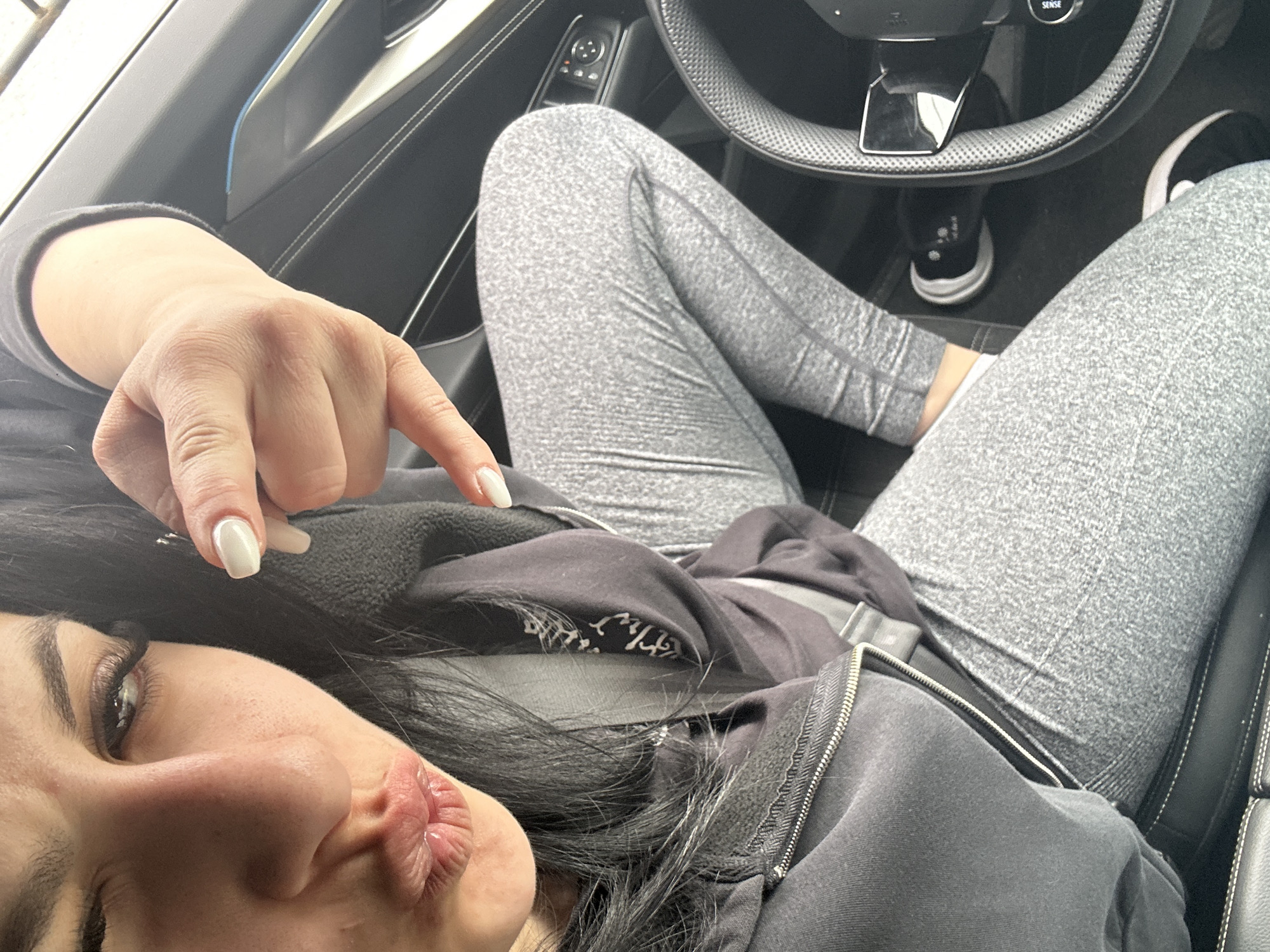 A person is reclining in the driver's seat of a car, with their legs resting on the steering wheel. They are wearing gray leggings and a black zip-up hoodie. The person's face is visible, and they have long dark hair and are pouting their lips. Their right hand is raised, with the index finger pointing downward. The background includes the car's interior, such as the dashboard and seats. The location is not discernible within the confines of the image.