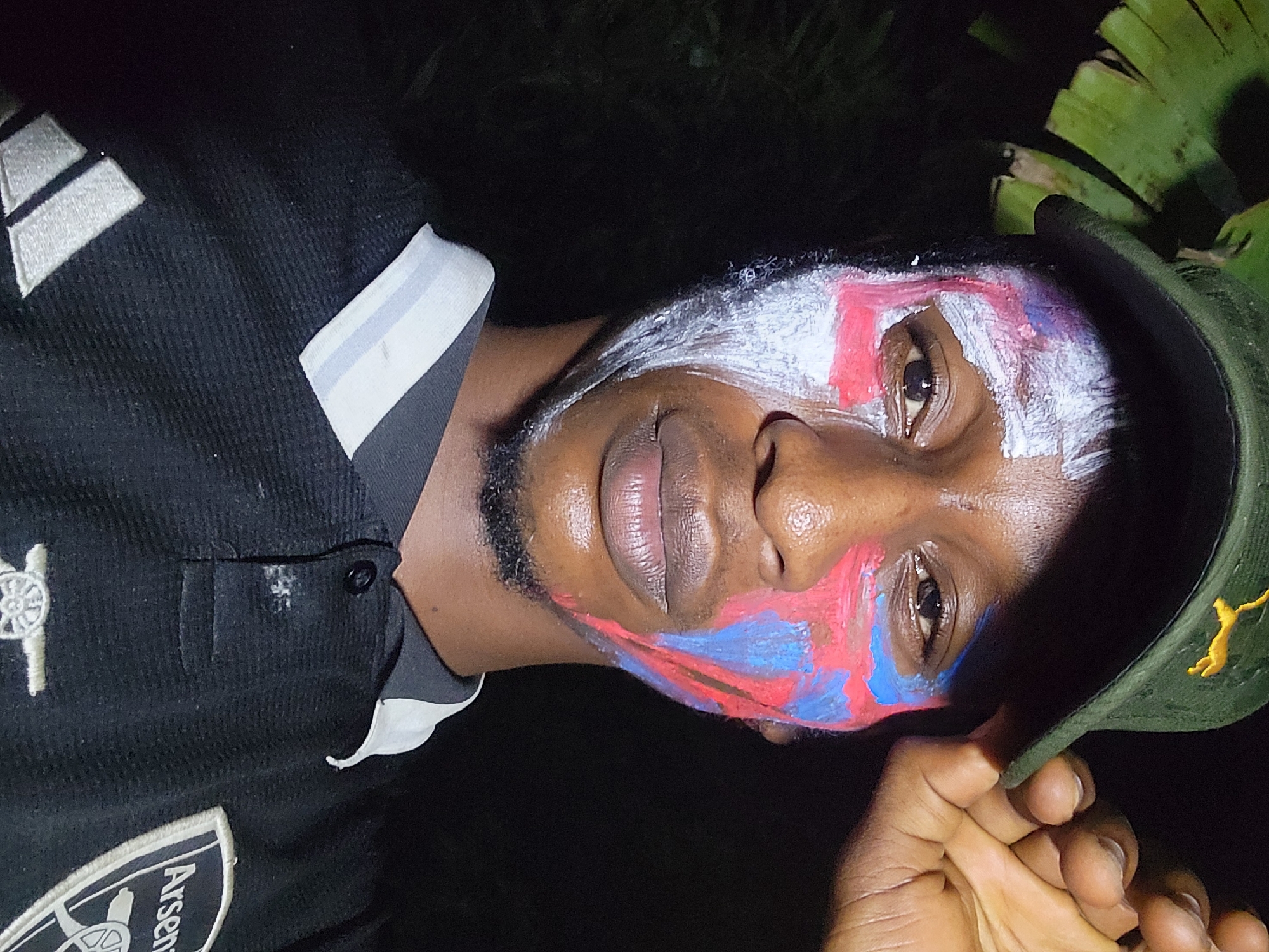 A close-up, low-angle shot shows a person with dark skin and face paint. The paint is white on the forehead, red on the upper cheek, and blue on the lower cheek. They are wearing a dark, corduroy shirt with a light gray collar and an Arsenal logo on the lower left. A green cap with a yellow logo is held in their hand. The background is dark, with some green leaves visible in the upper right. The location is Unknown, Unknown.