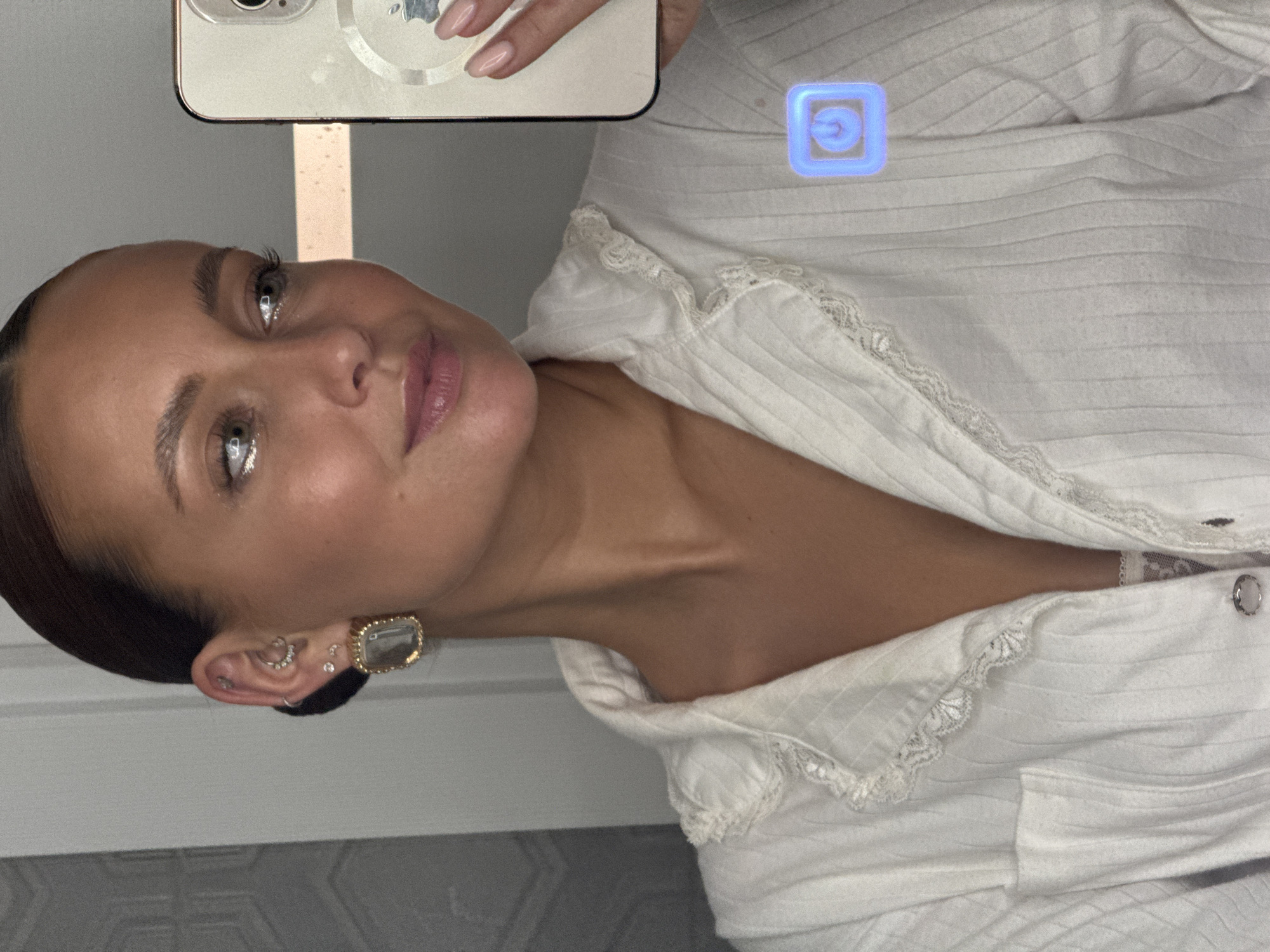 A woman with dark hair pulled back, wearing a white top with lace detailing, is taking a selfie with a white smartphone. The phone partially obscures the upper portion of her face. She is wearing multiple earrings in her left ear, including a large, square, gold-toned earring. A blue square with a power symbol is superimposed on the upper right portion of the image. The background appears to be a room with a gray and white patterned wall and a vertical metallic strip. There is no information in the image to indicate the location is Chaikovsky, Russia.